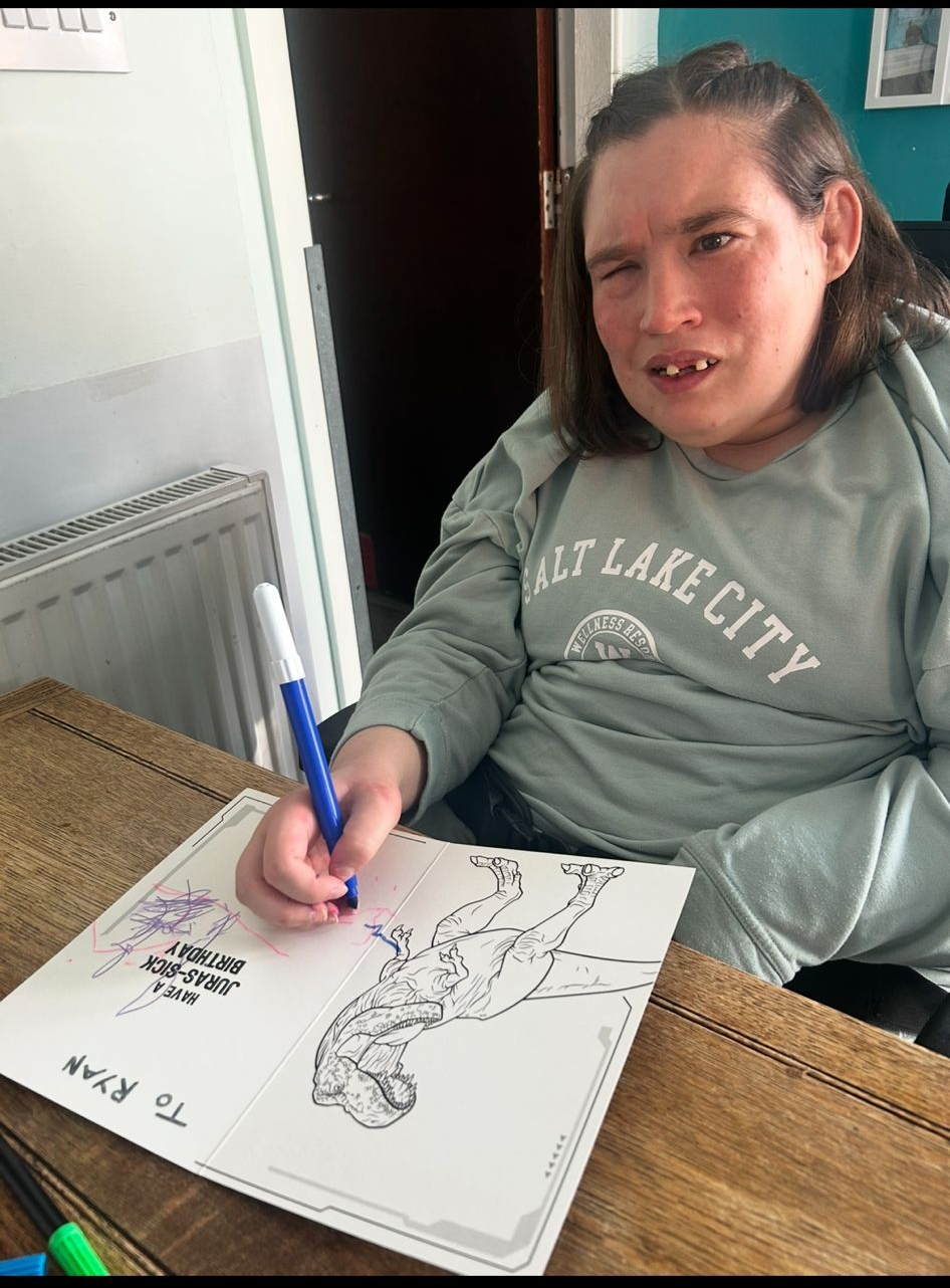 Person with long hair seated, drawing with blue pen on paper, on a wooden table. Wearing a grey sweatshirt.