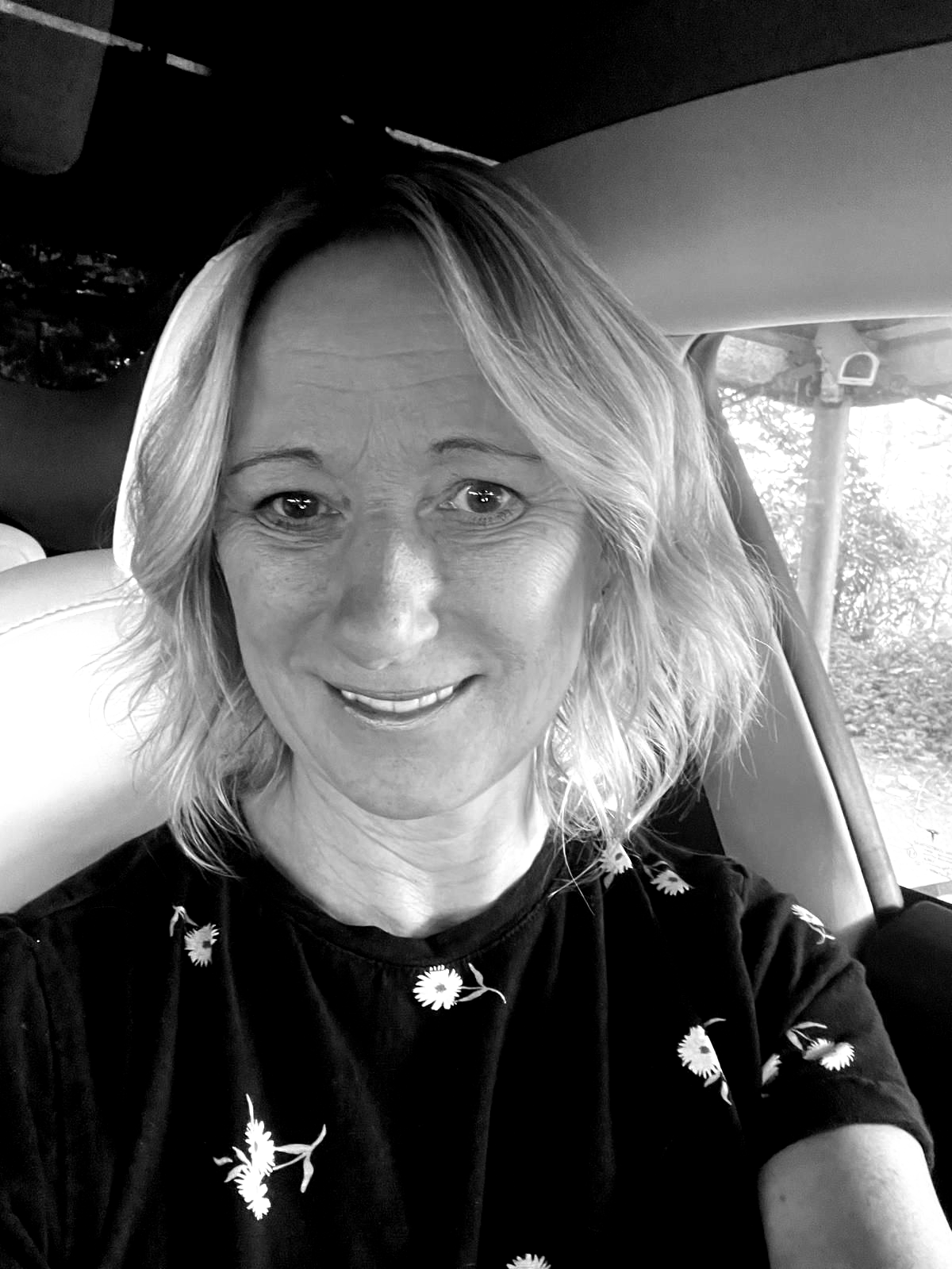 Woman with blonde hair smiling in a car, wearing a dark shirt with a floral pattern.