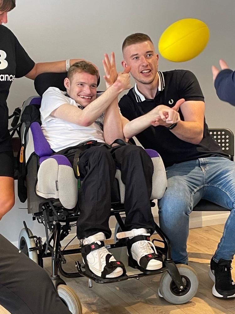 Two men in a room, one in a wheelchair, are playing catch with a yellow rugby ball.