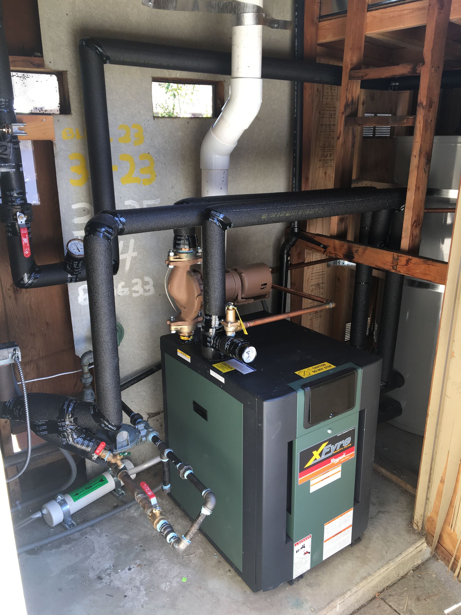 A boiler is sitting in a room with pipes and a ladder.