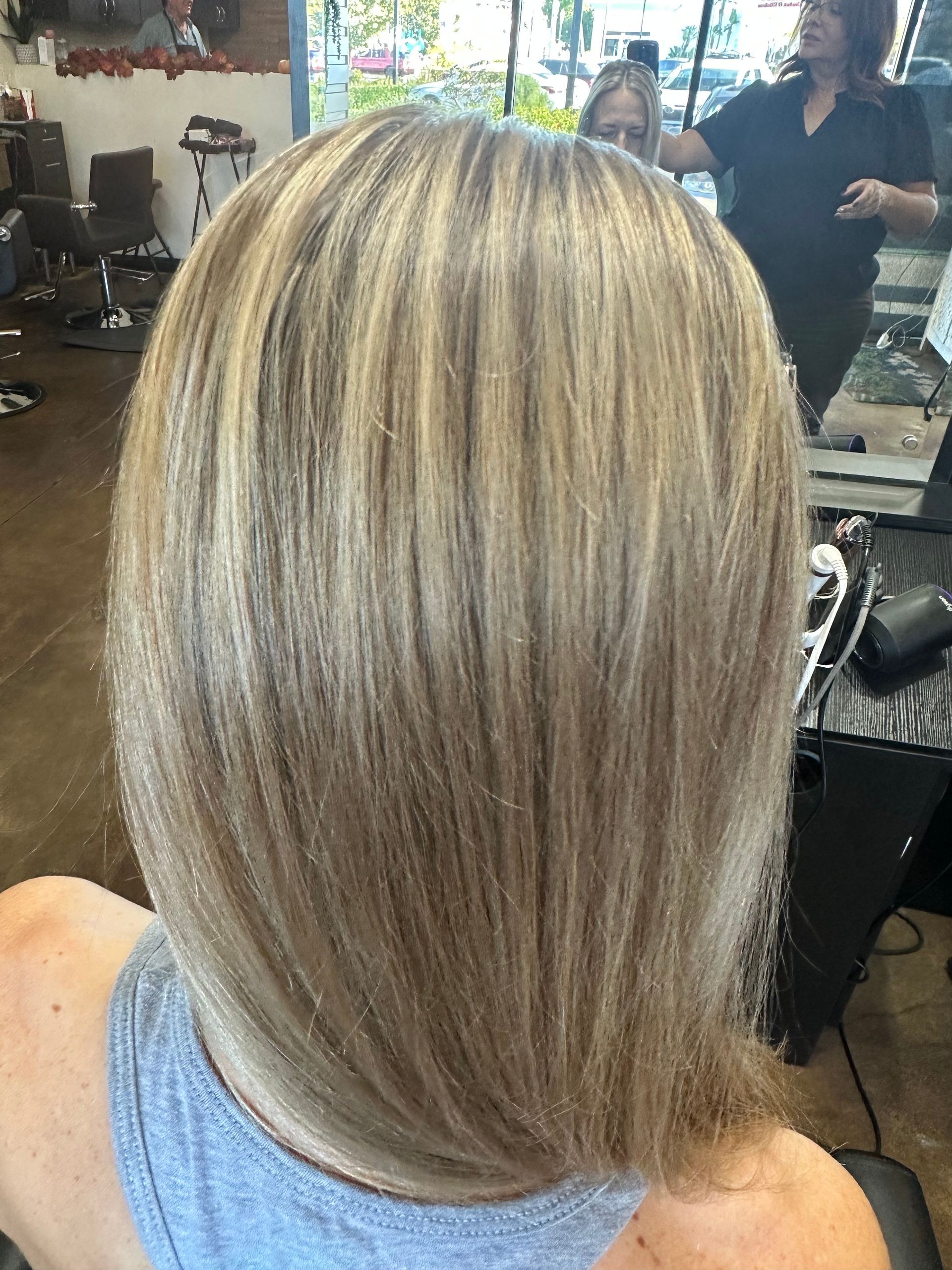 Blonde hair with highlights; person seated in a salon; stylist in background.