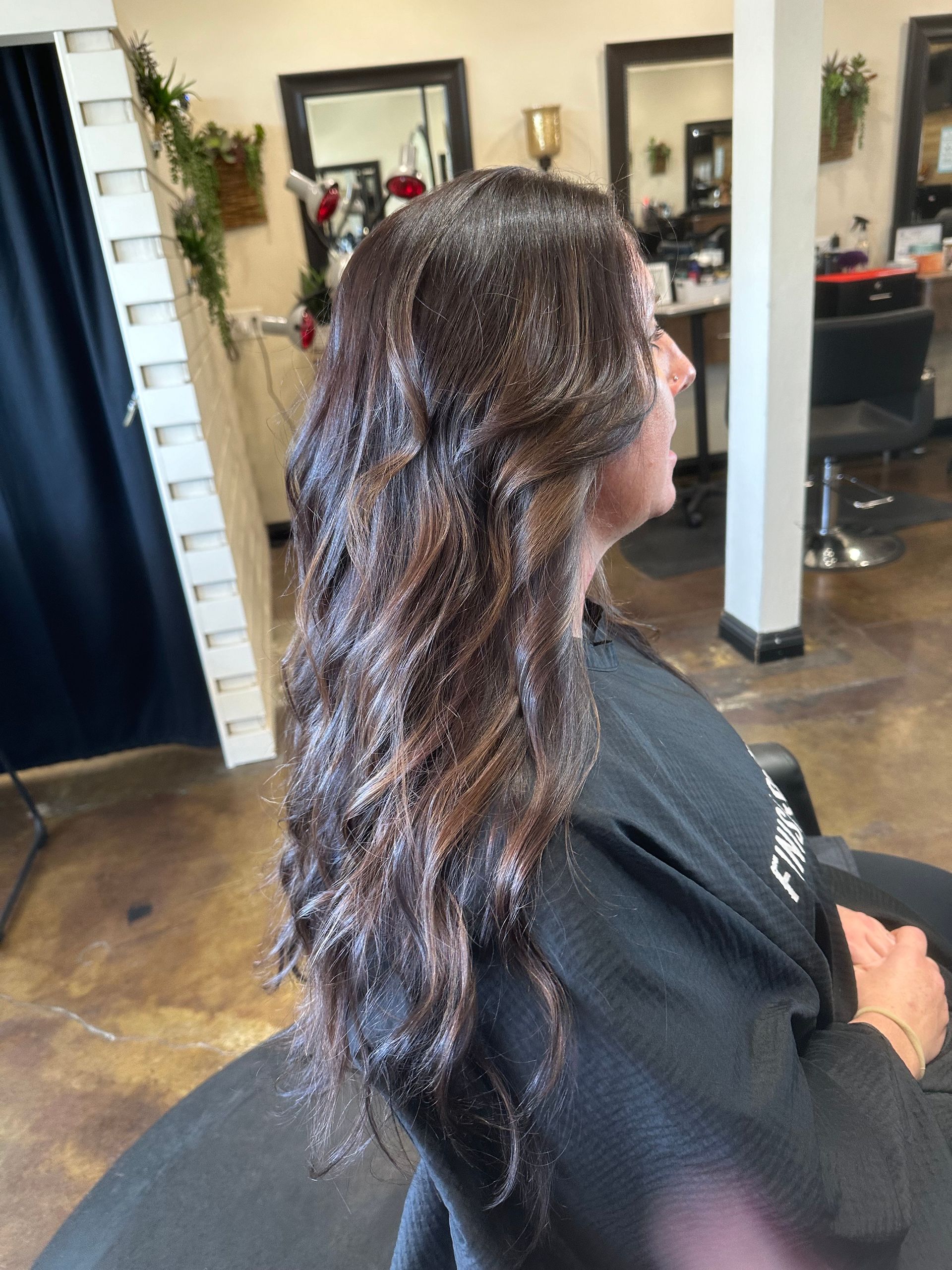 Woman with wavy dark brown hair in a salon chair, wearing a black cape, with mirrors and decor in background.