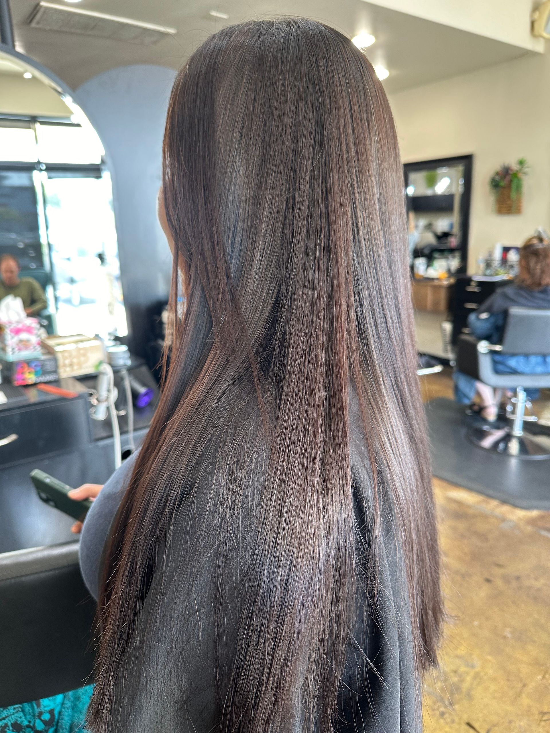 Woman with long, sleek dark brown hair in a salon, seen from the back.