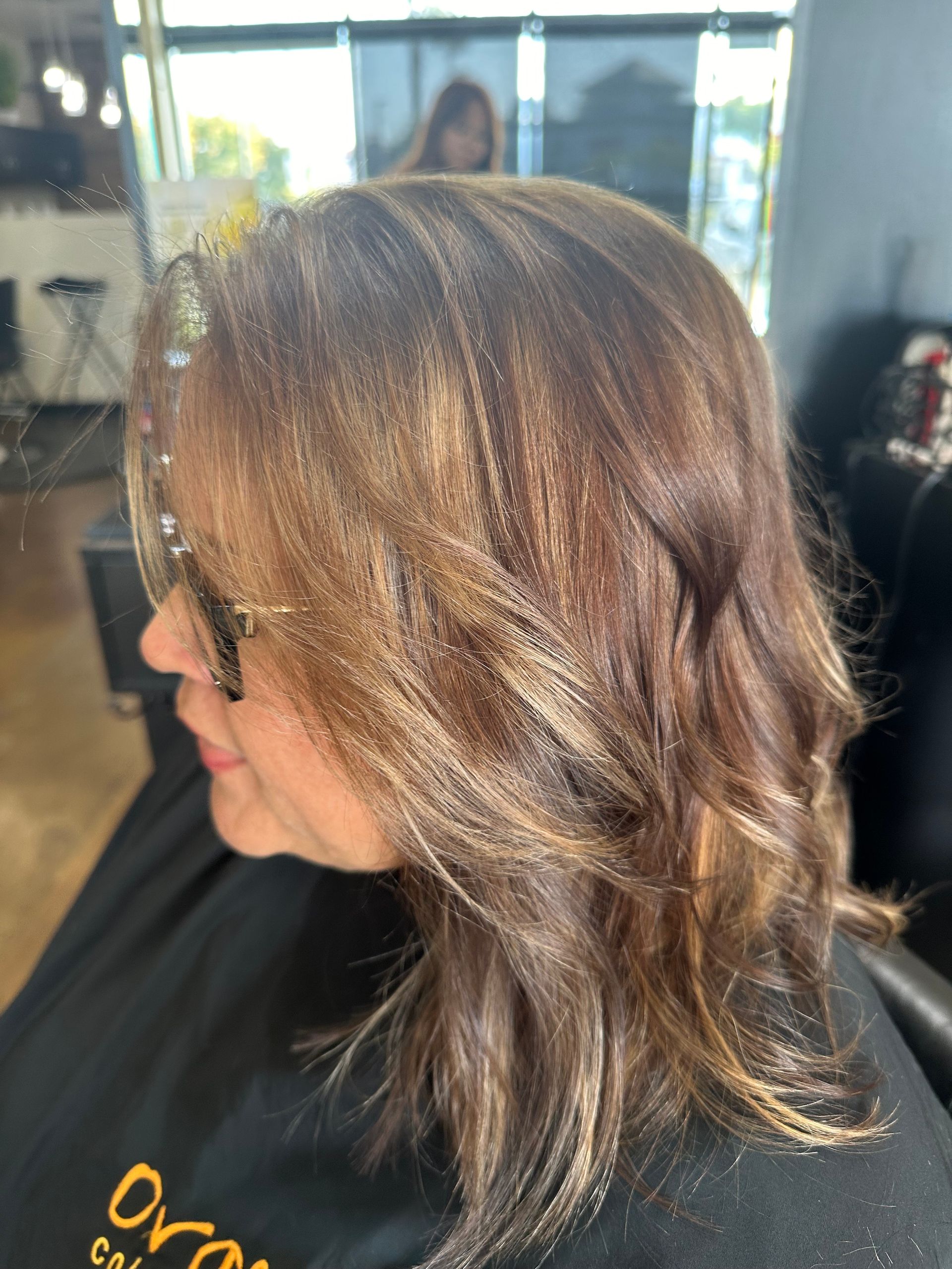 Woman with brown hair and blonde highlights, wearing sunglasses, in a salon.