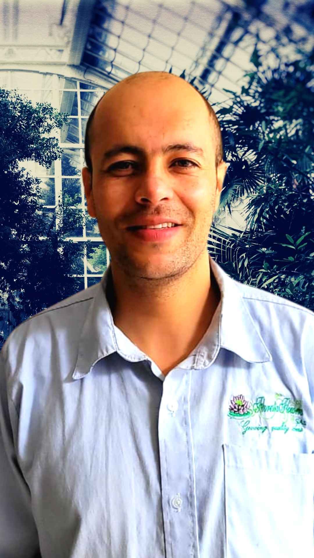 un hombre calvo con una camisa azul sonríe frente a un invernadero.