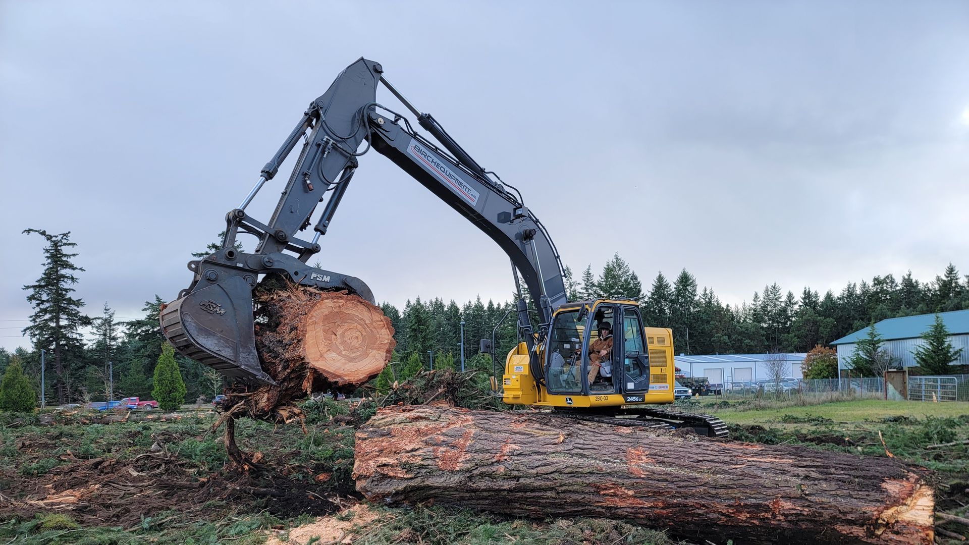 Excavator lifting a large tree trunk, outdoors. Yellow and gray machine, green field, trees and buildings in the background.