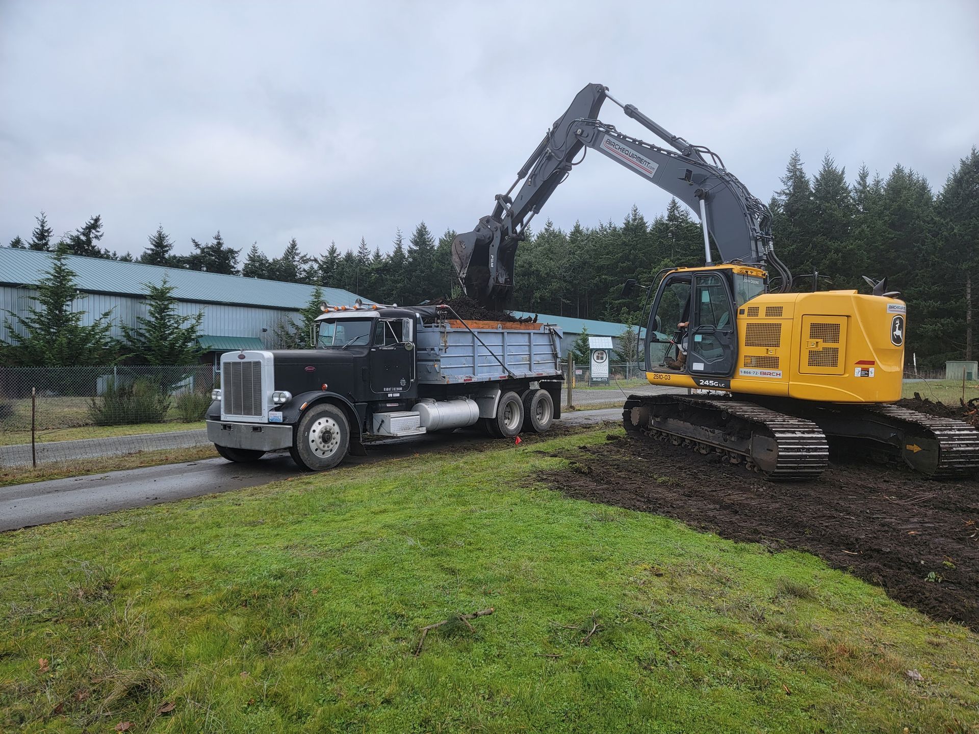 A John Deere excavator loading dirt into a black dump truck on a muddy, grassy site.