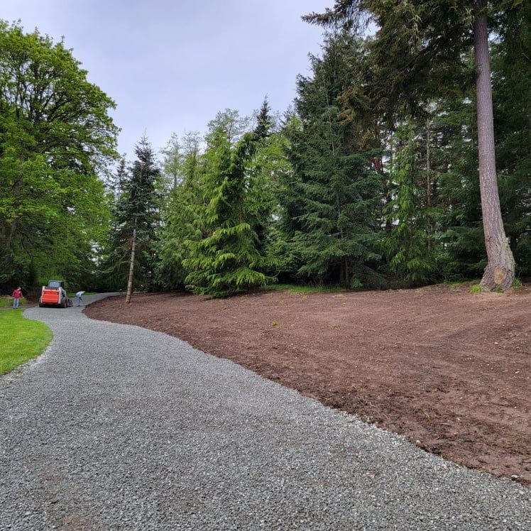 Gravel driveway curves past a freshly tilled area next to a forest. A small orange construction vehicle is visible.