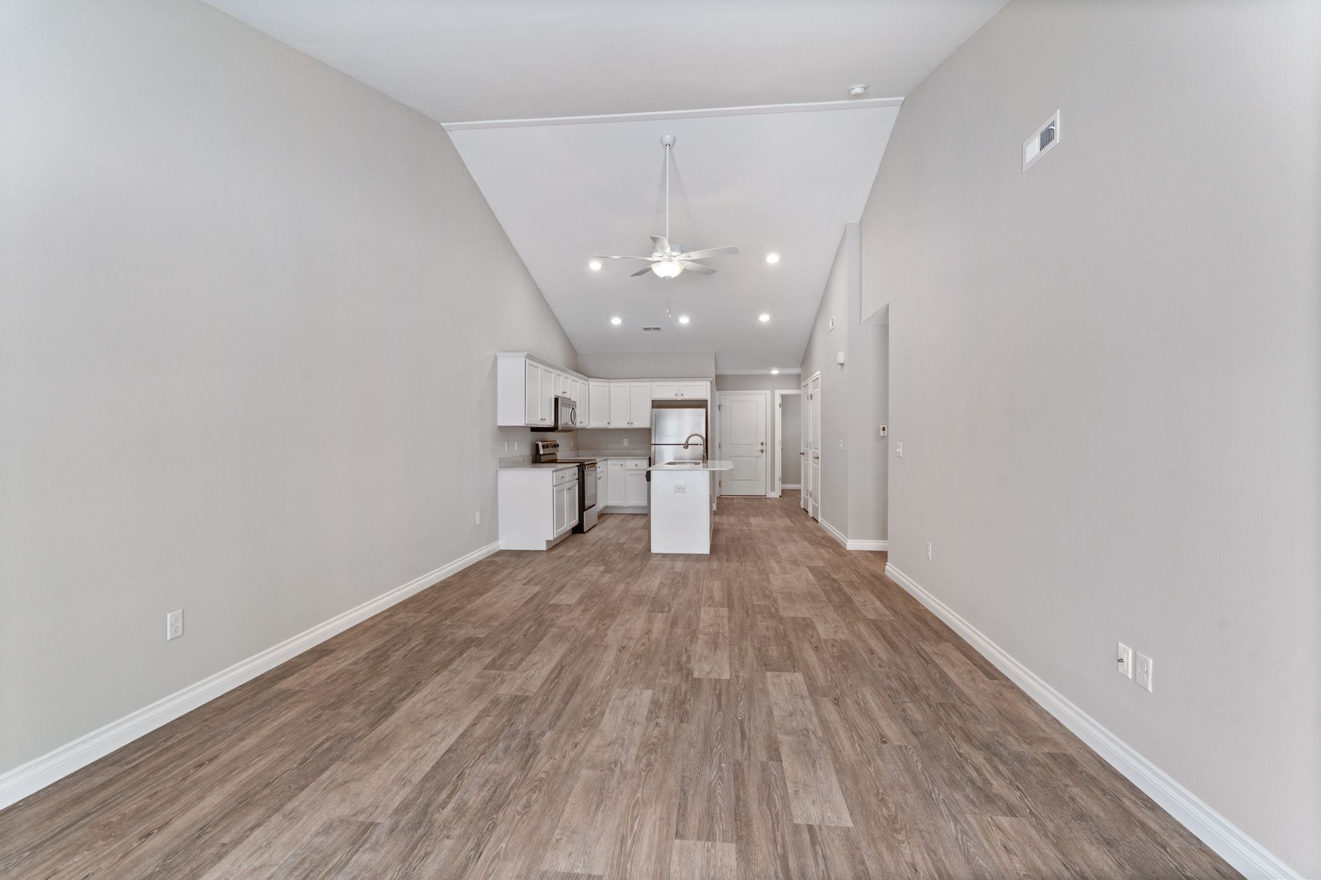 Empty, open-plan interior with kitchen in the background, light wood flooring, and white walls.