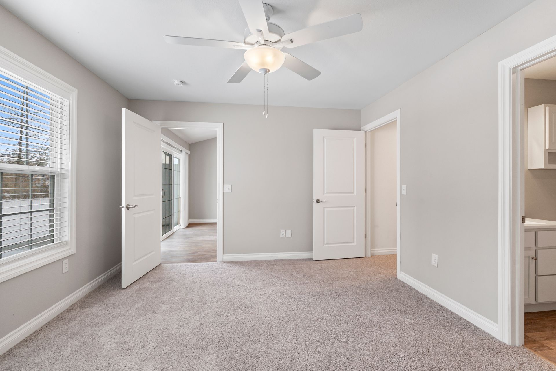 Empty bedroom with light gray walls, carpet, and three white doors; window with blinds; ceiling fan.