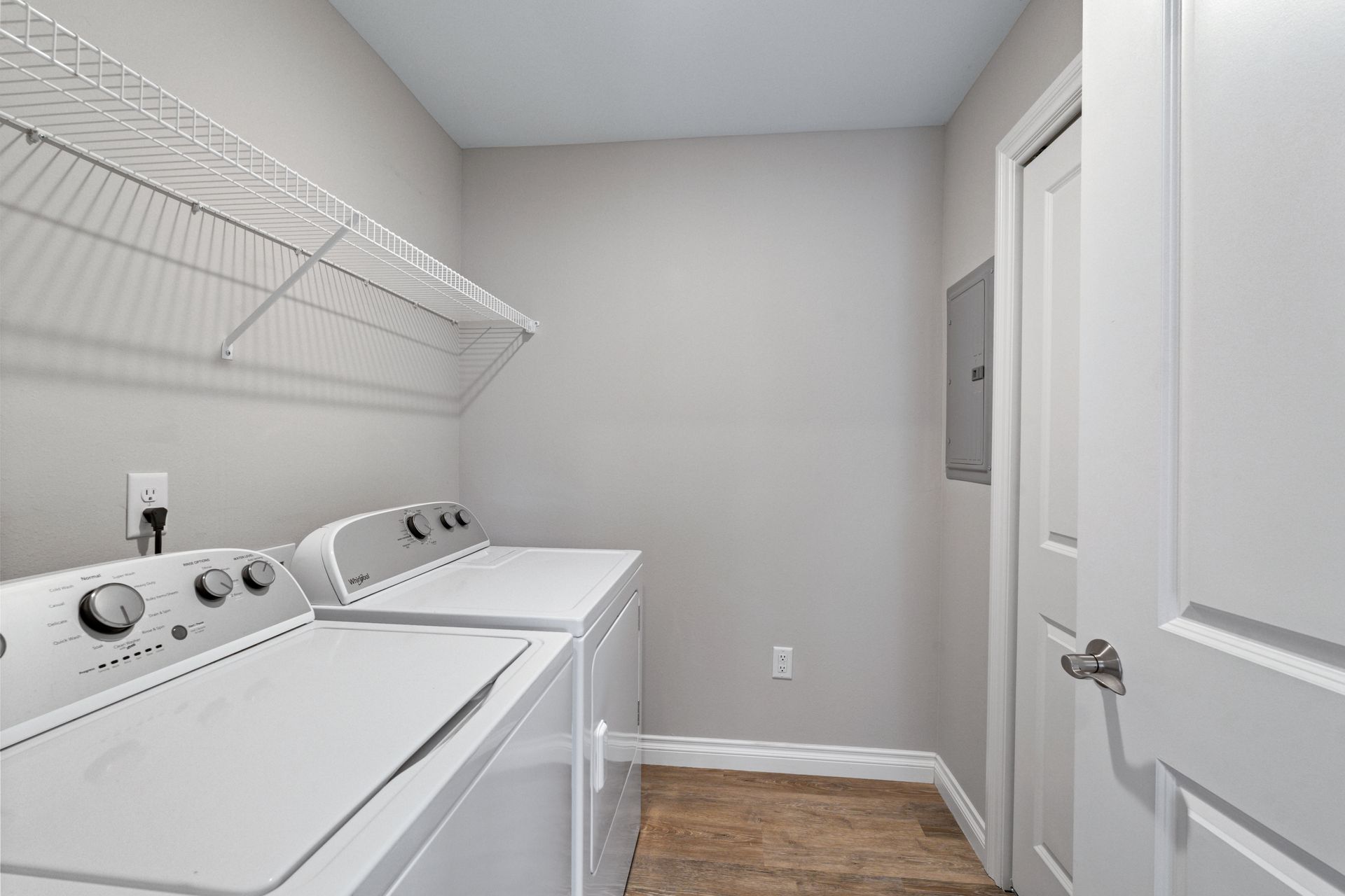 Laundry room with white washer, dryer, wire shelf, closed door, and electrical panel on gray wall.