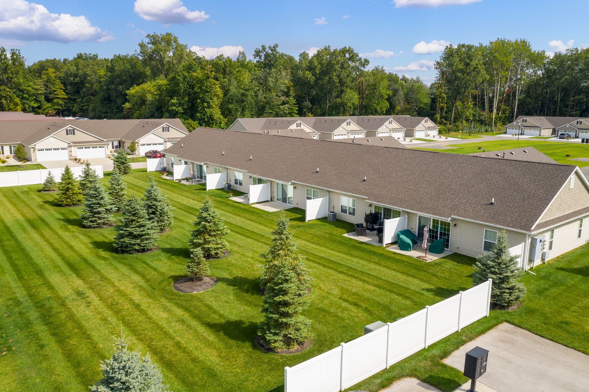 Row of white townhouses with brown roofs and green lawns, trees in the yard.