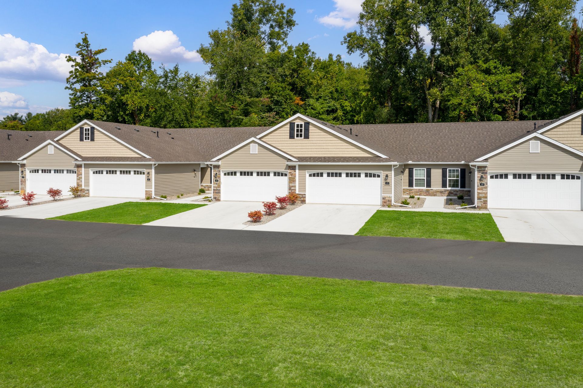 Row of attached tan single-story houses with white garage doors, asphalt driveway, green lawn, and trees in the background.