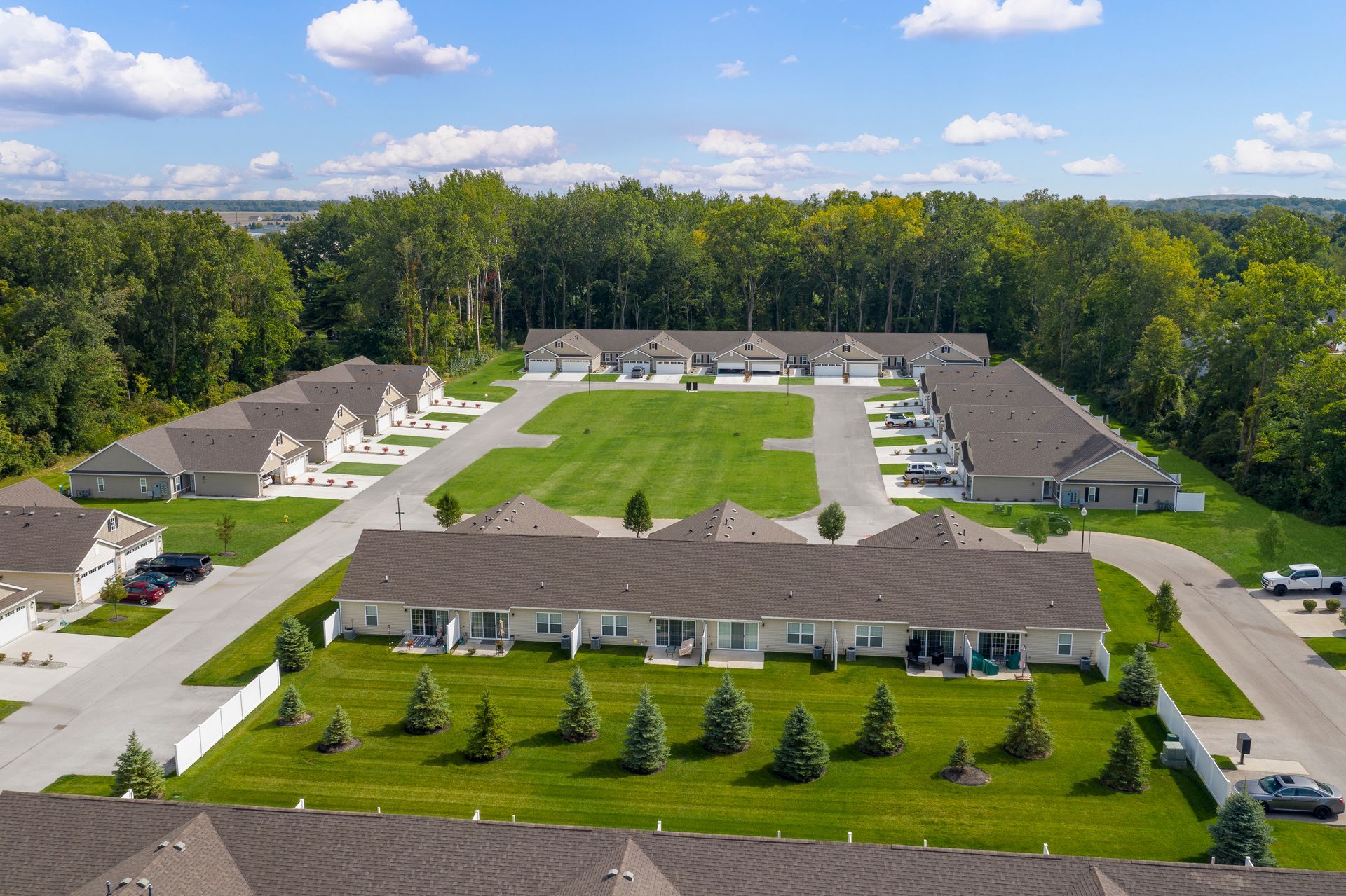 Aerial view of multiple one-story townhomes with green lawns and trees. Blue sky.