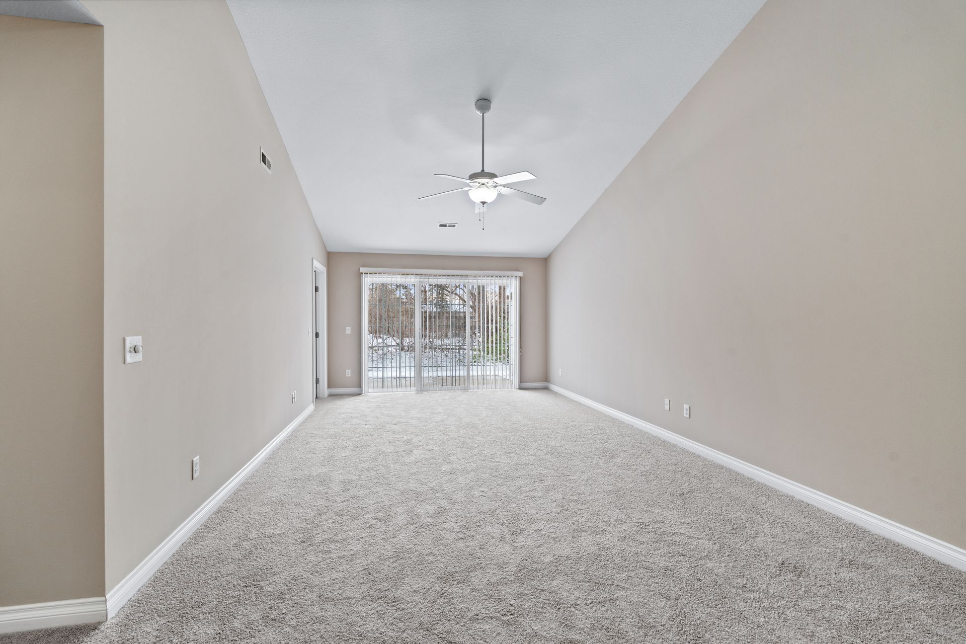 Empty room with beige walls, light carpet, ceiling fan, and sliding glass door to a snowy exterior.