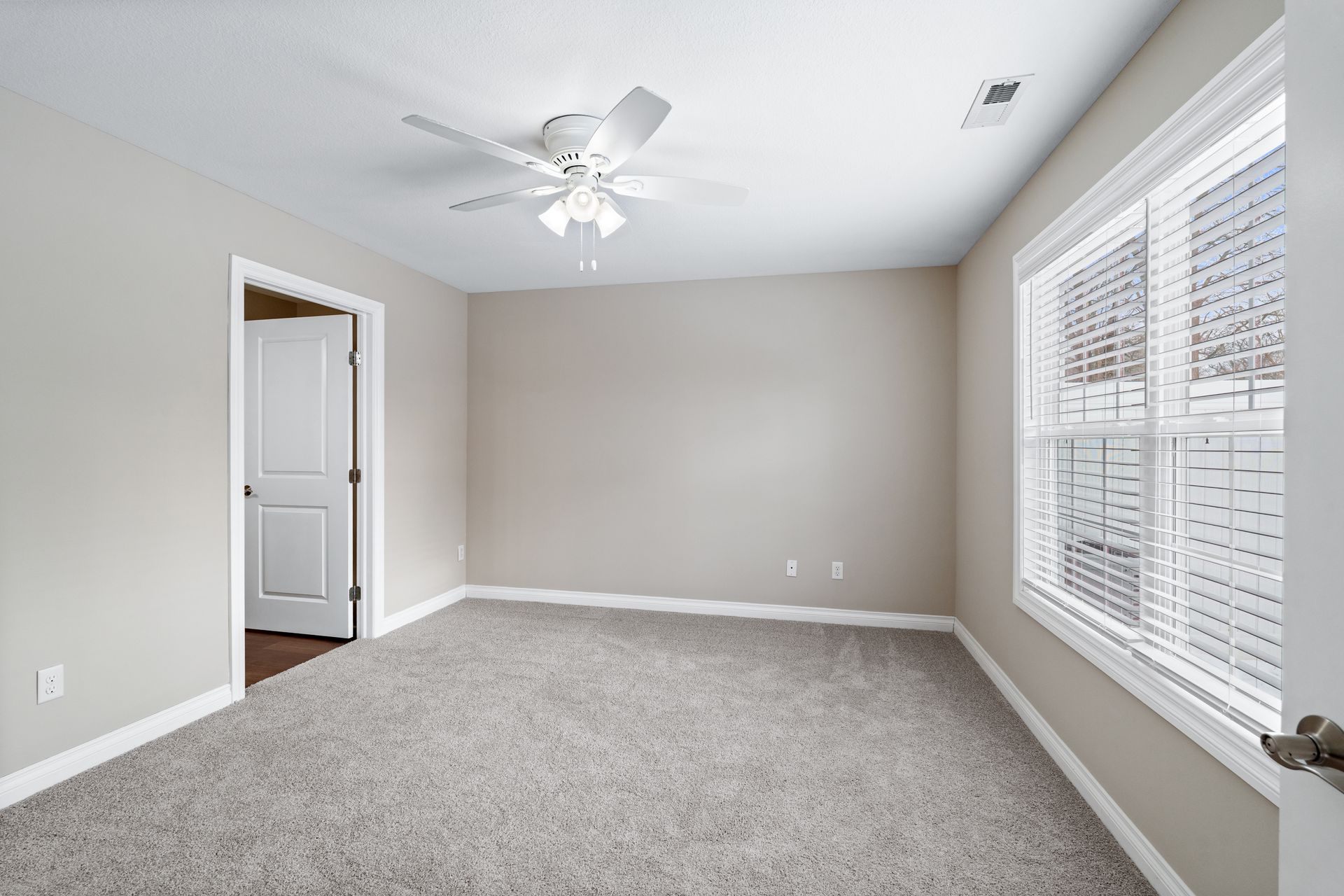 Empty room with beige walls, gray carpet, white ceiling fan and trim, closed white door, and window with blinds.
