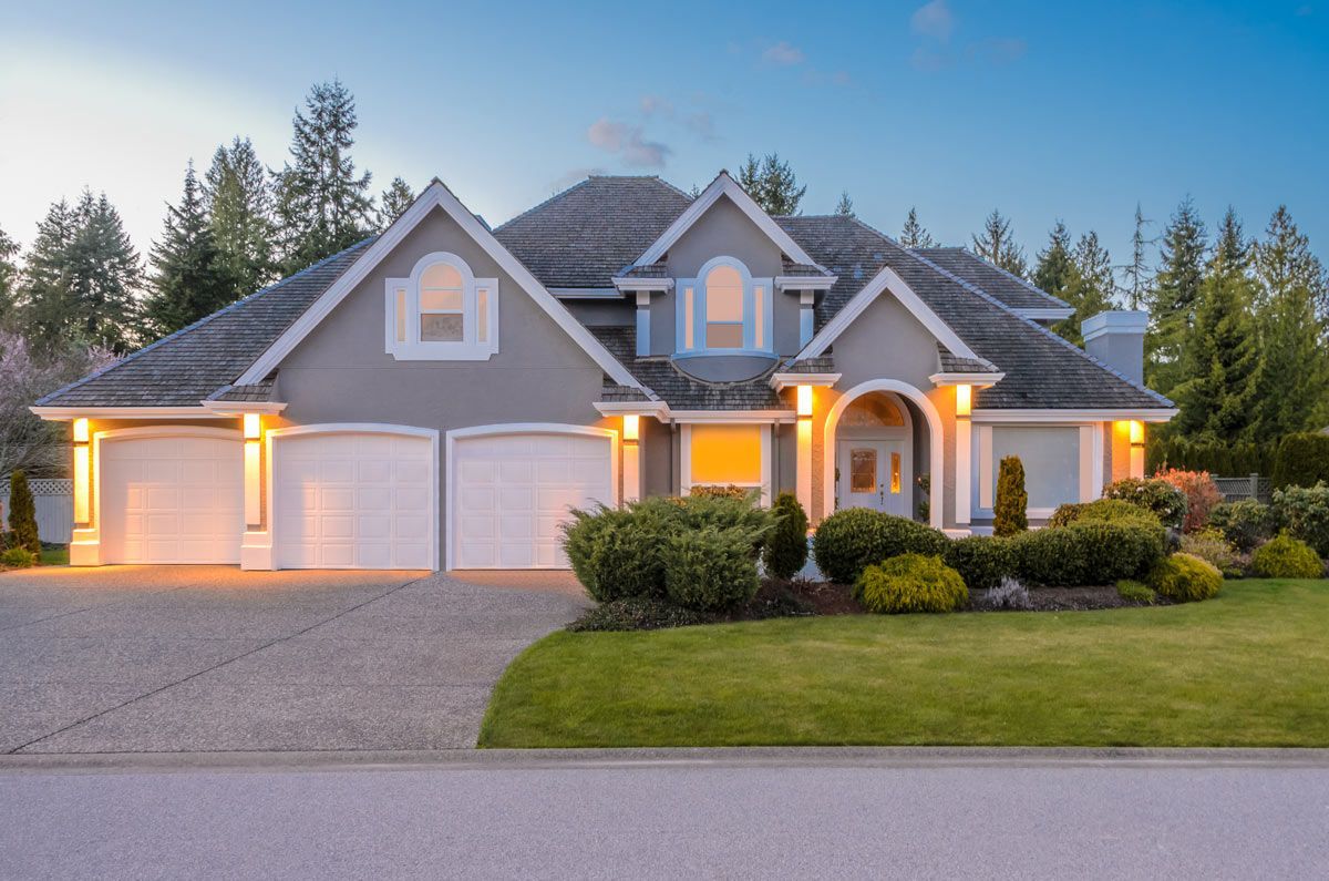 Gray house with white garage doors, lawn, and driveway; evergreen trees in the background.