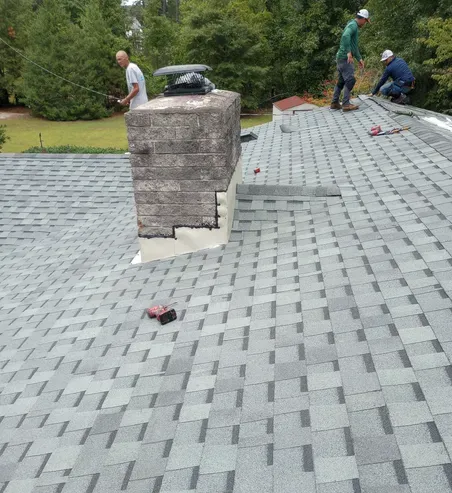 Three roofers installing shingles around a chimney on a gray-shingled roof in a yard.