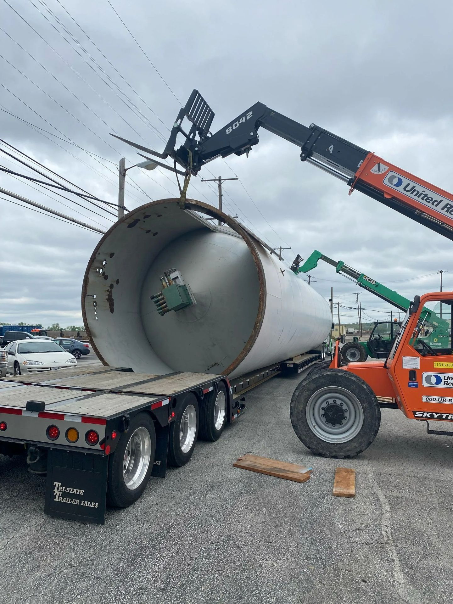 A large industrial tank is lifted by a forklift from a flatbed truck in an outdoor setting.