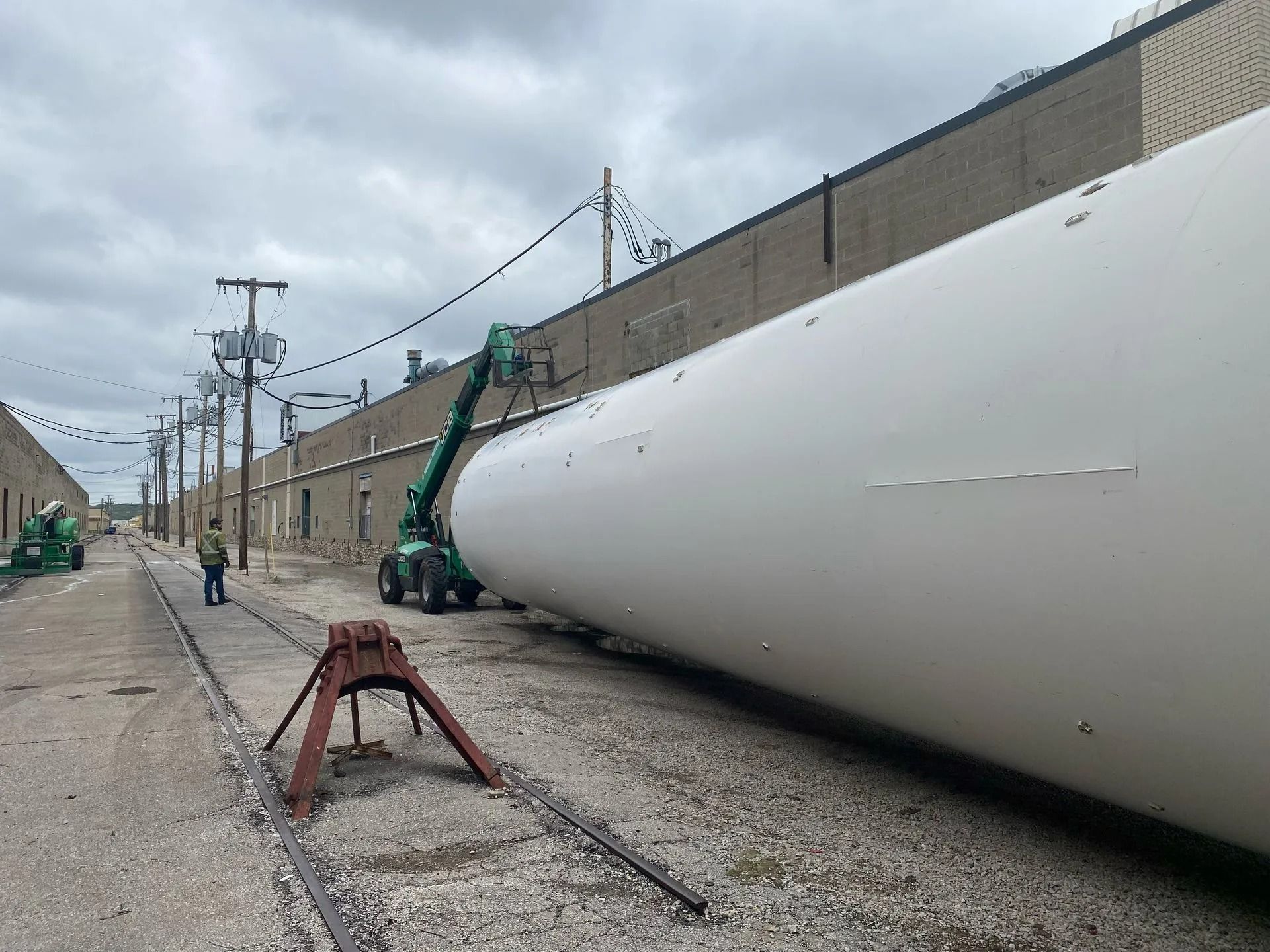 A large white cylindrical object lies on railroad tracks, being handled by a green crane. A person stands nearby.