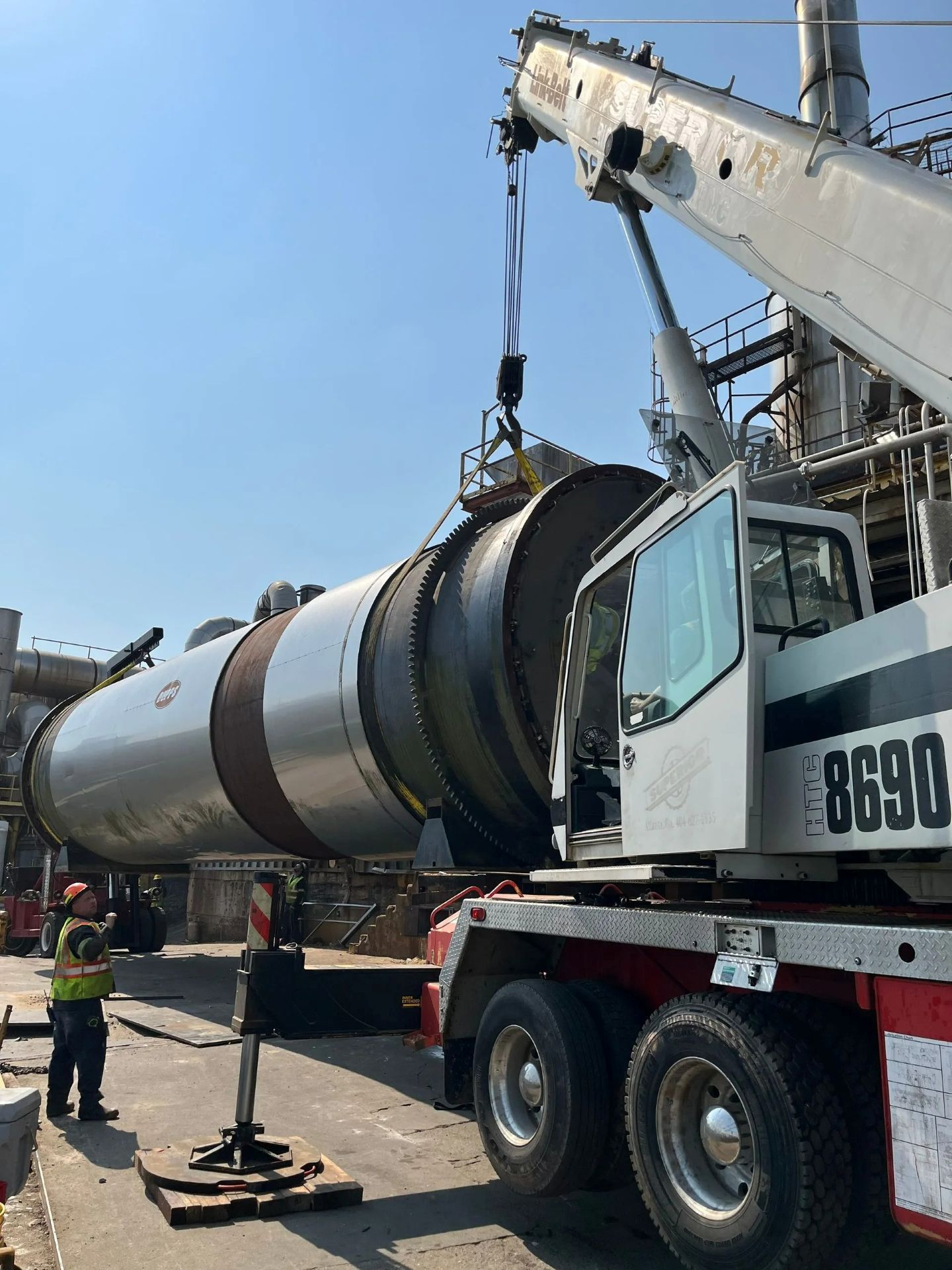 A crane lifts a large industrial cylindrical tank at a plant, with a worker observing.