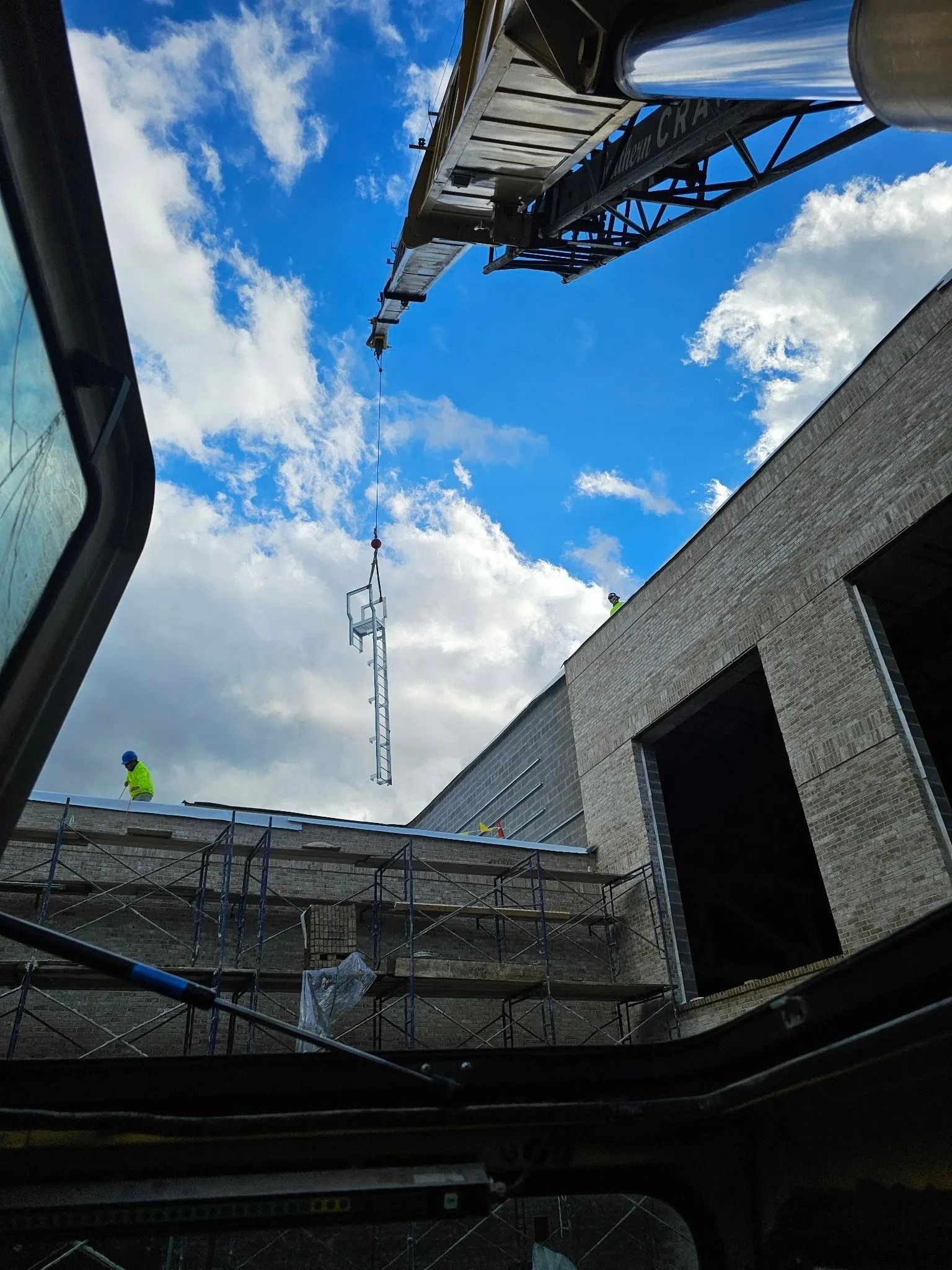 Construction site with crane lifting a steel structure towards a building against a cloudy blue sky.