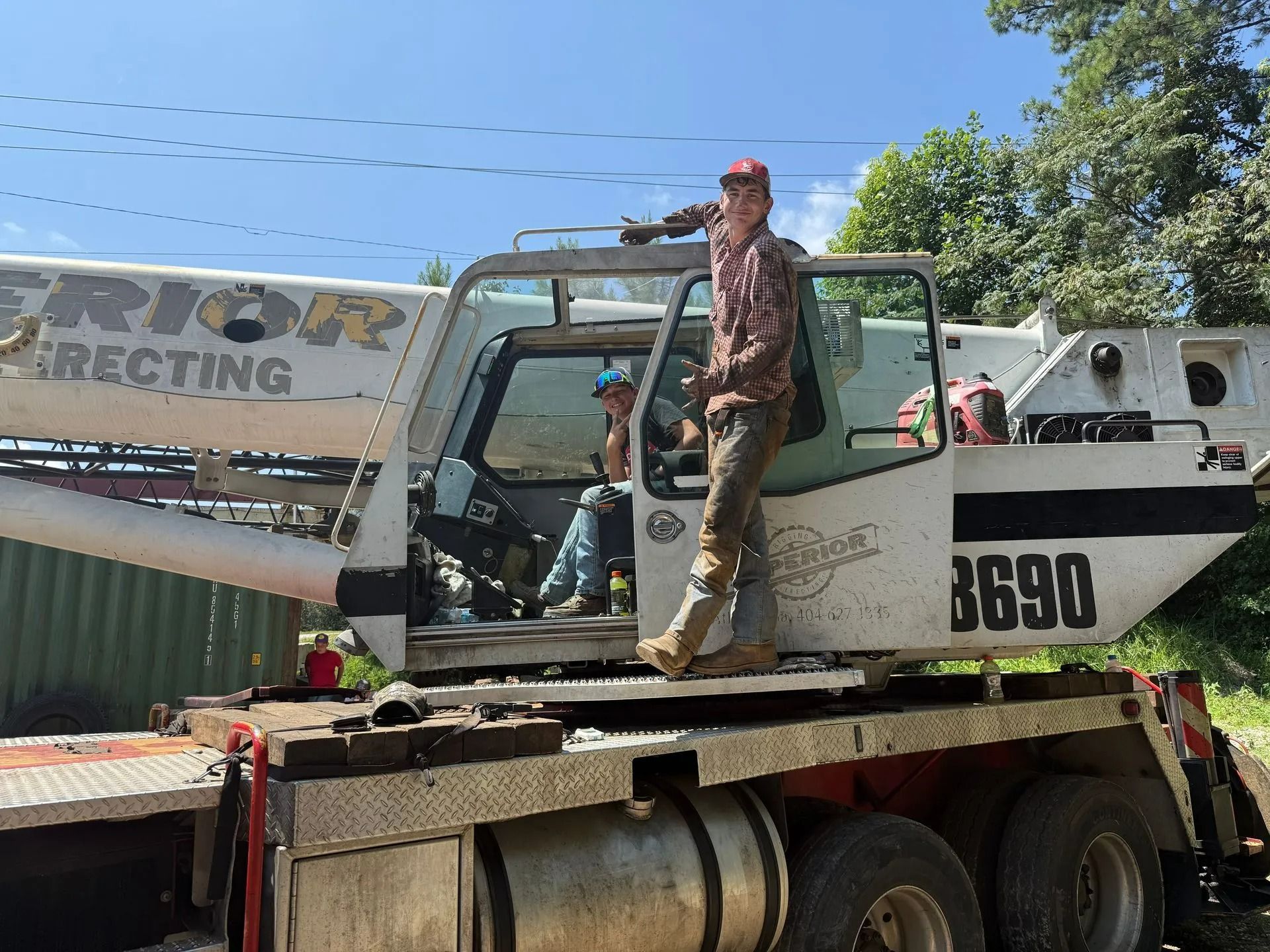 Two people near a crane truck. One stands in the doorway, the other sits in the cab. Sunny, outdoor setting.
