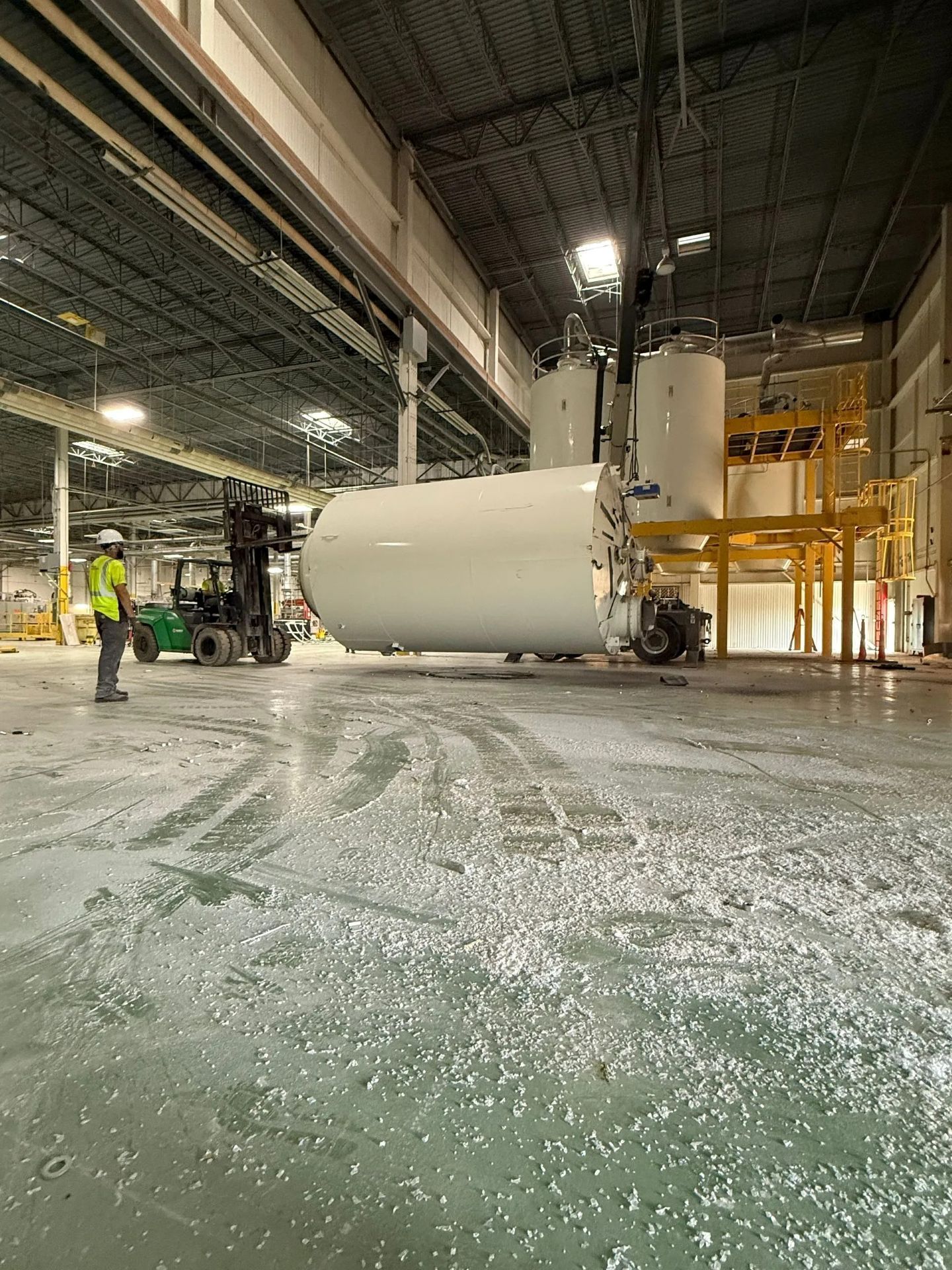 Forklift moving large white tank inside a factory. Worker in safety vest watches.