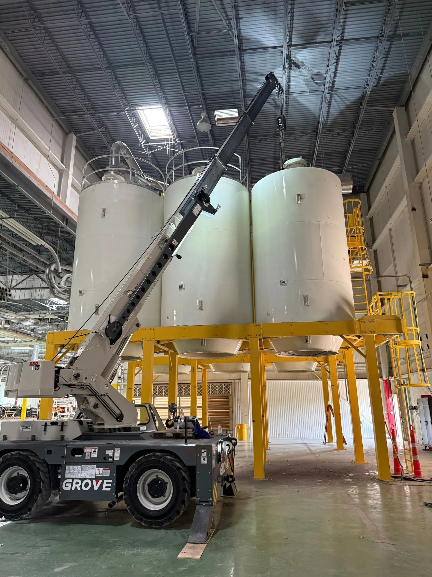 A Grove crane lifting in an industrial setting with three large white tanks on a yellow platform.