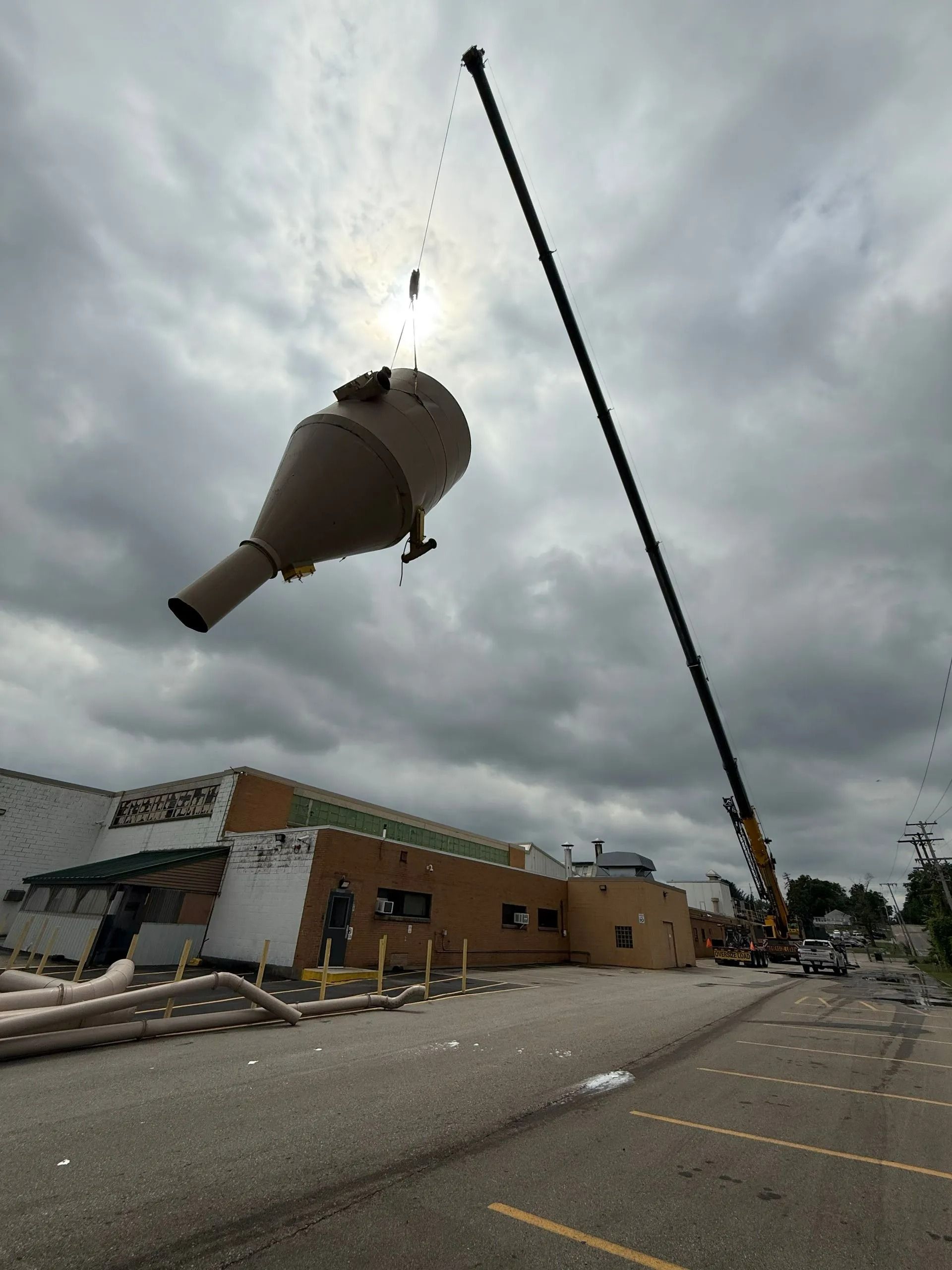 A crane lifts a large brown industrial cone over a building. Overcast sky.