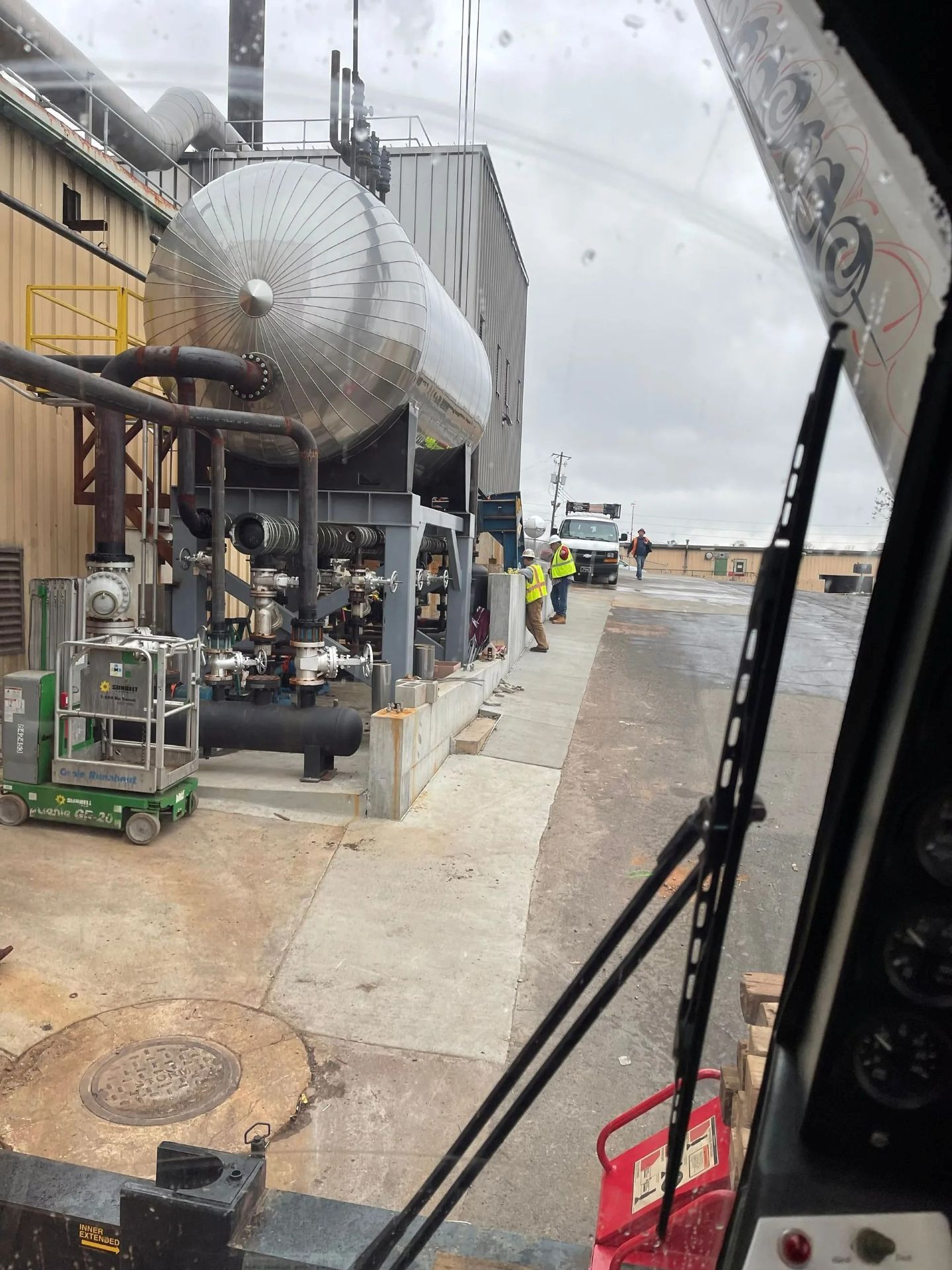 Industrial tanks and machinery outside a building, viewed from a vehicle. Workers near a truck.