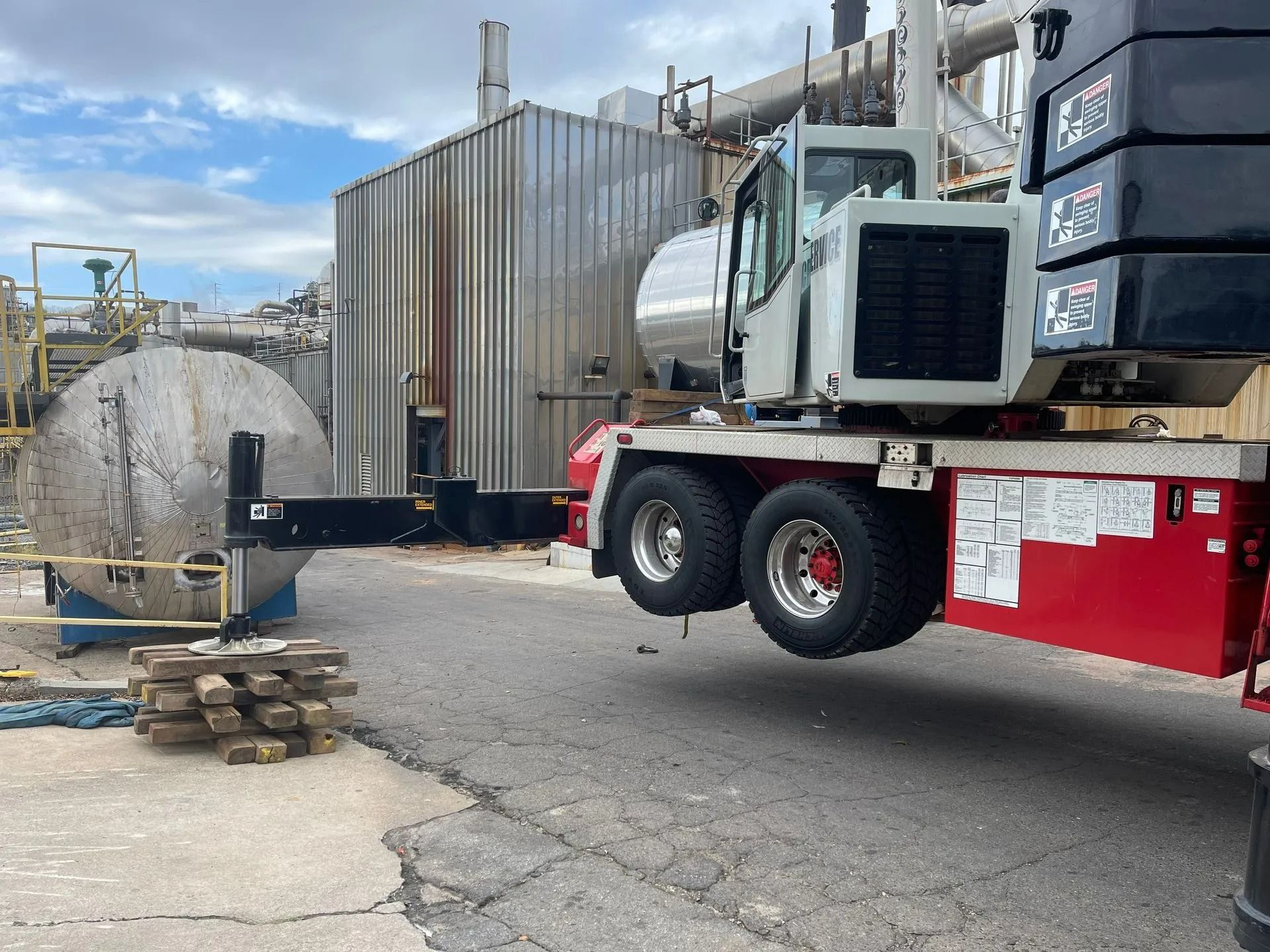 A mobile crane lifts a large, cylindrical metal object near a metal industrial building.