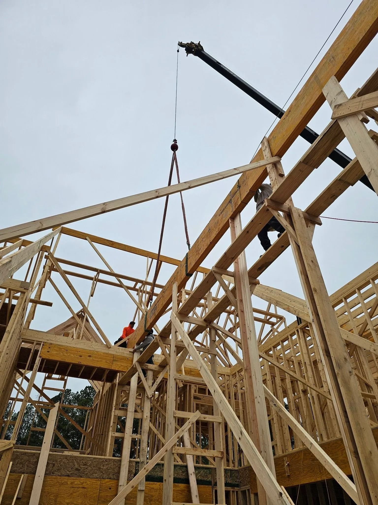 Construction workers use a crane to lift a wooden beam onto a partially built house frame. Cloudy sky.