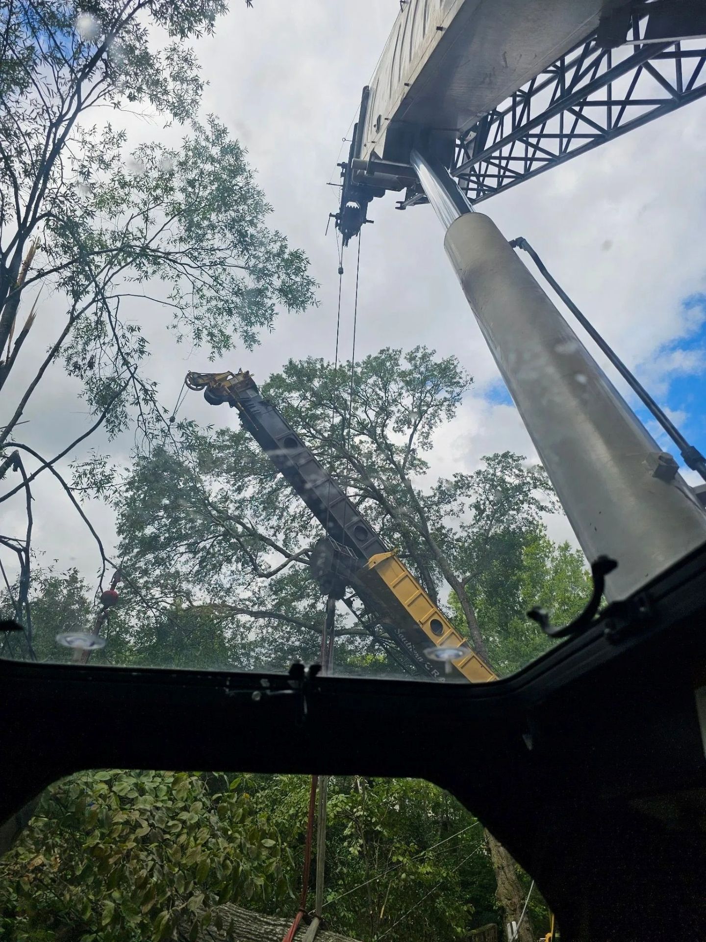 View from inside a crane cab, looking up at its boom extension against a cloudy sky and trees.