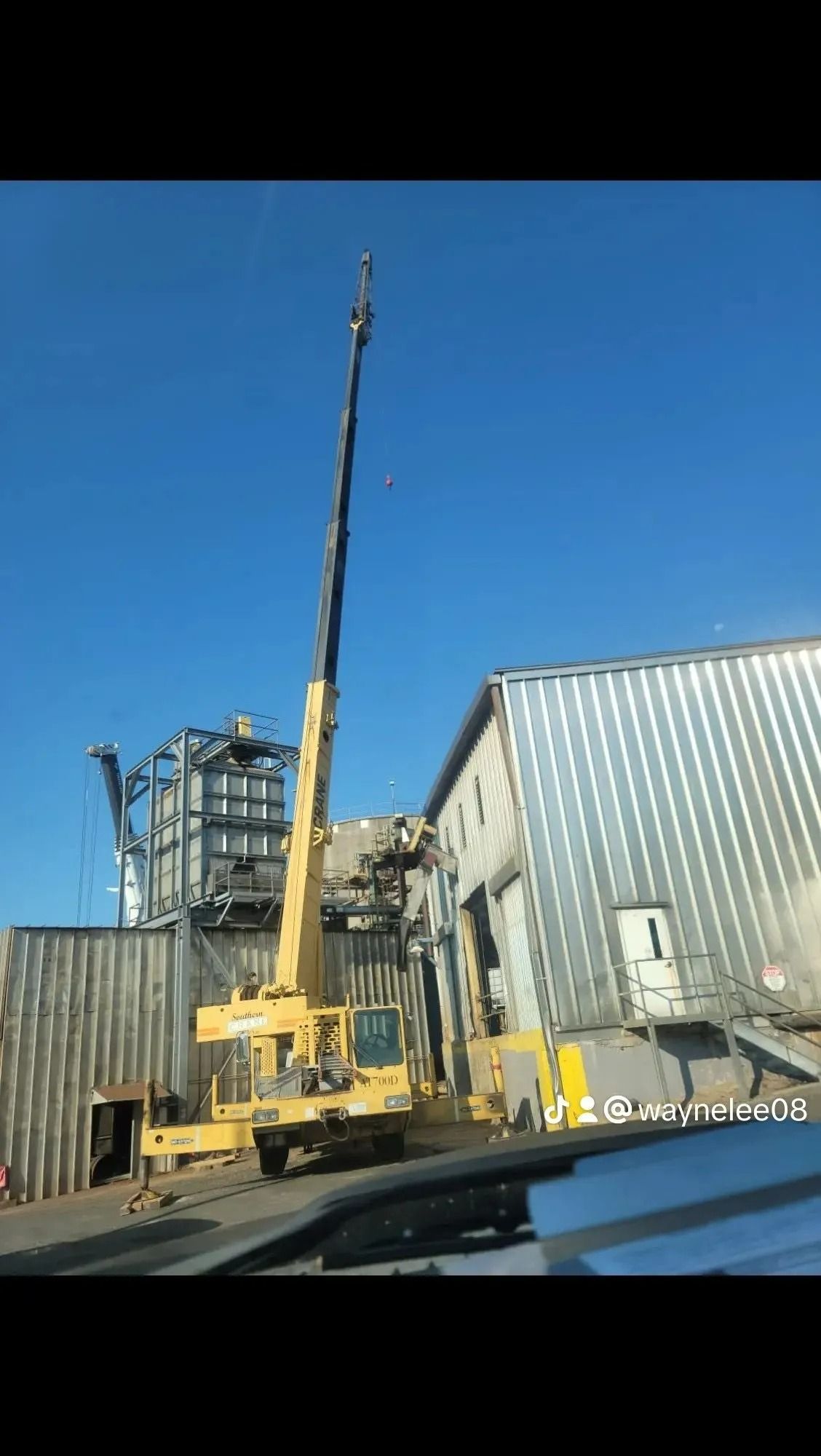 Yellow crane lifting near a metal building and structure under a clear blue sky.