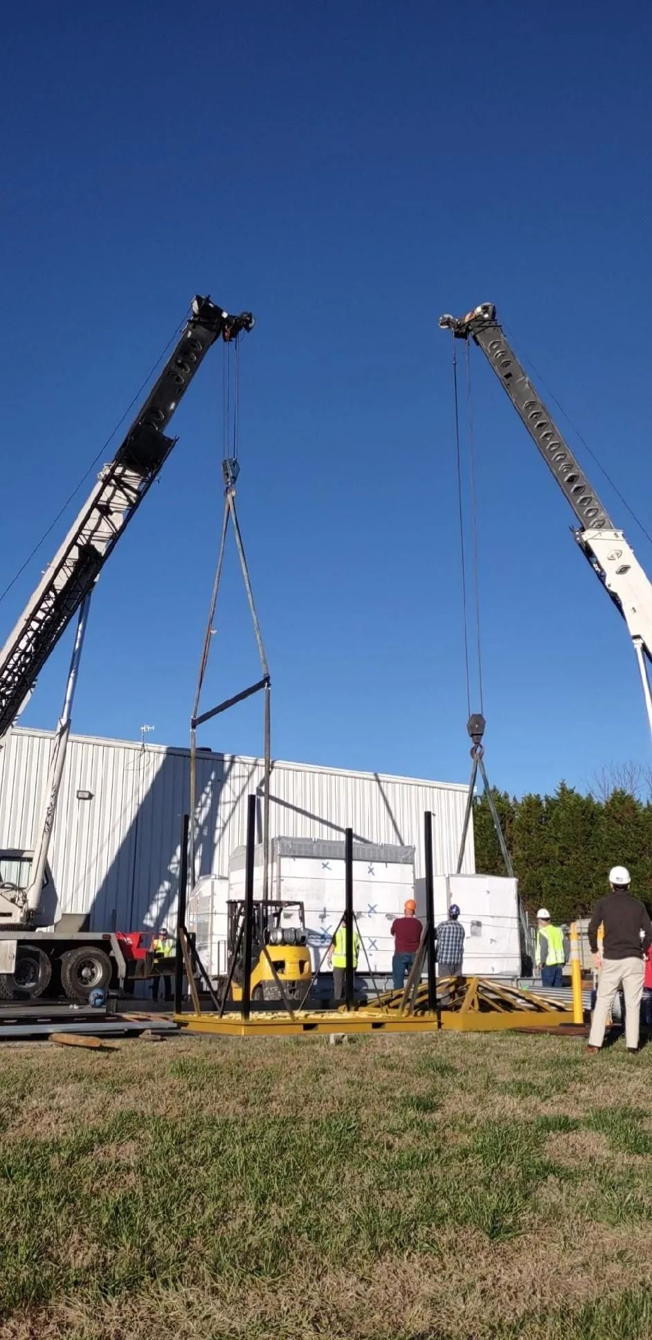 Two cranes lifting metal structures near a building, construction site on a sunny day.
