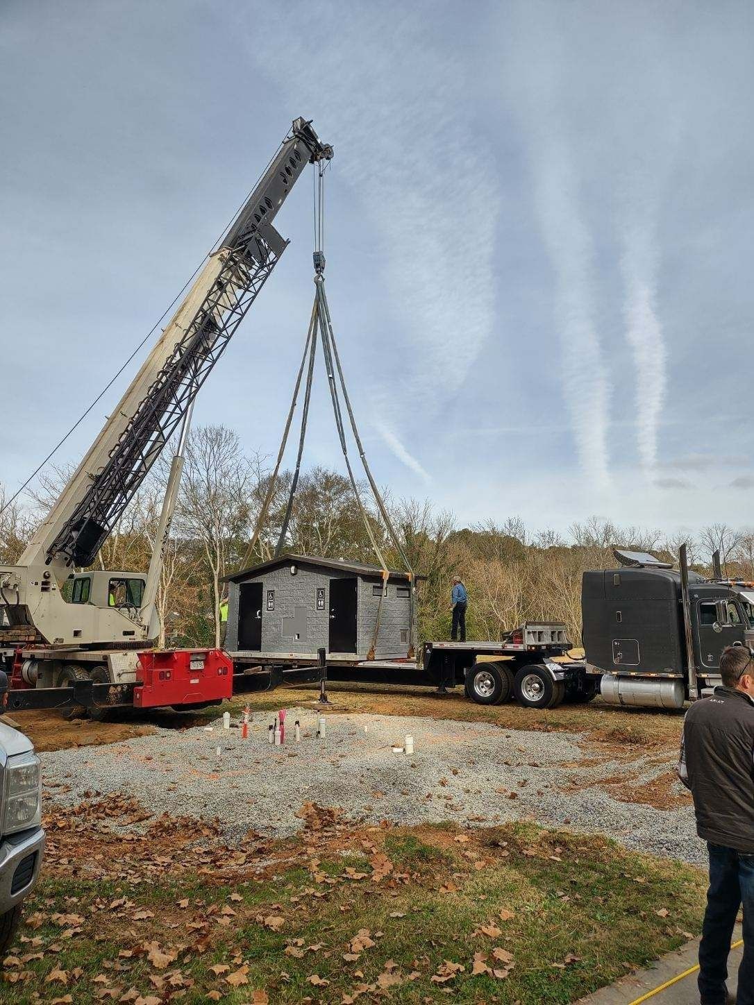 Crane lifting a gray building from a flatbed truck. Outdoors, construction site.