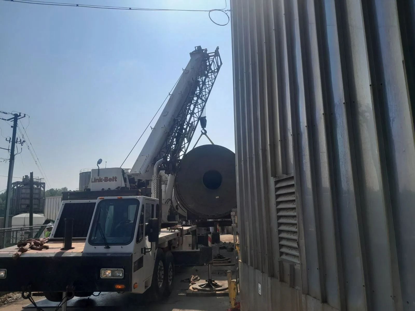 Crane lifting a large, cylindrical object near a corrugated metal structure on a sunny day.