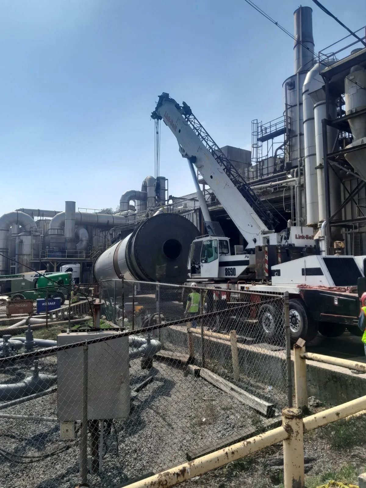 Crane lifting large cylindrical object at an industrial facility with piping and structures; sunny day.