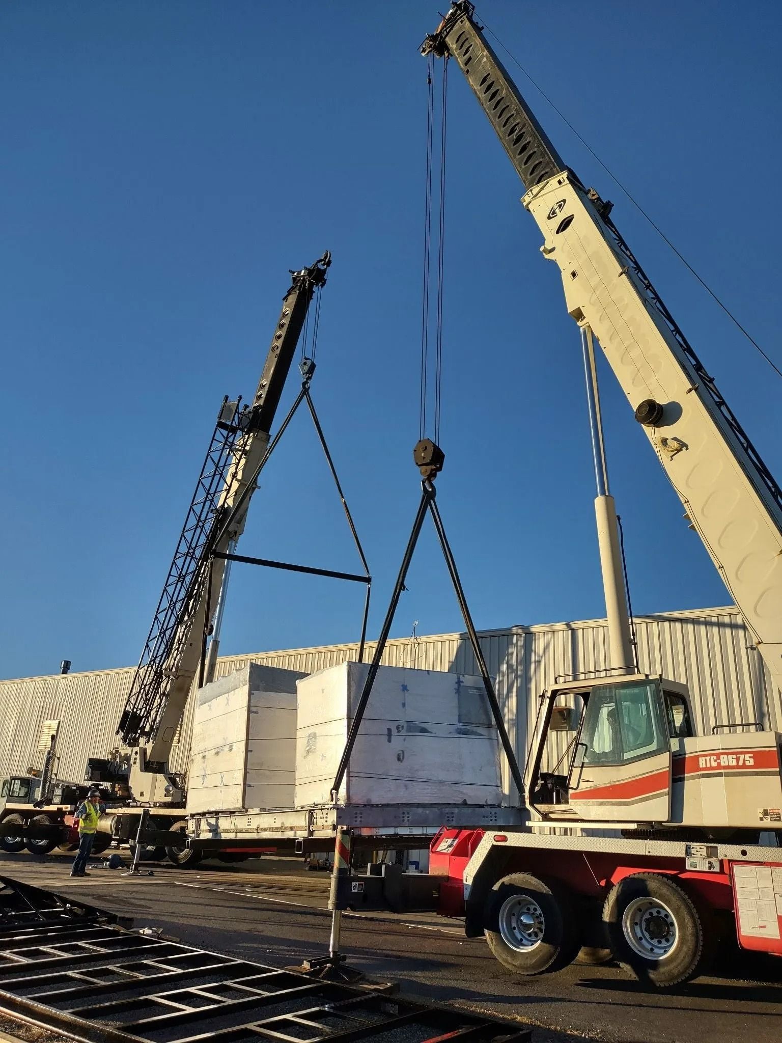 Two cranes lifting a large, rectangular object from a flatbed truck against a clear, blue sky.