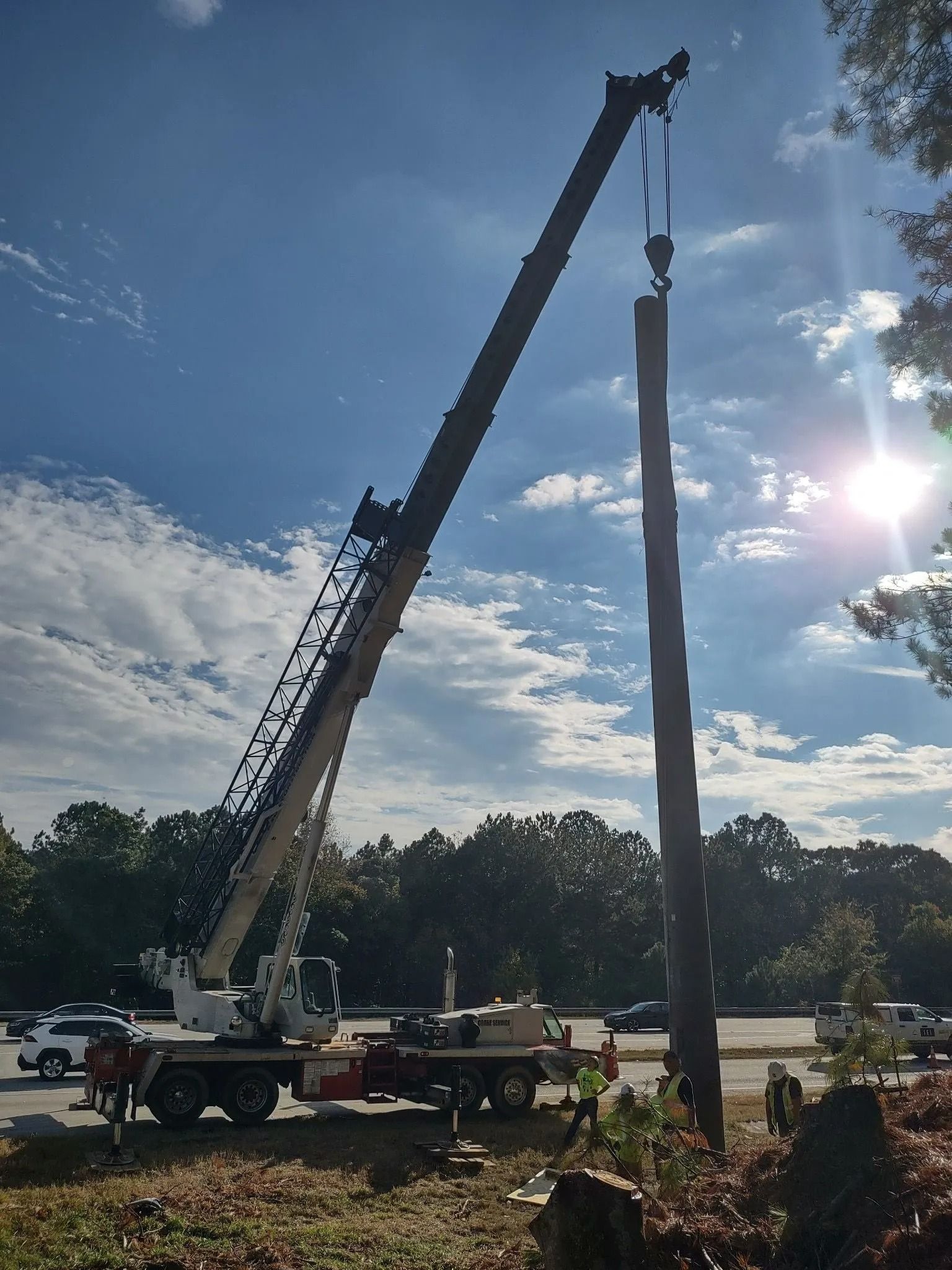 A crane lifting a tall, wooden pole against a partly cloudy sky.