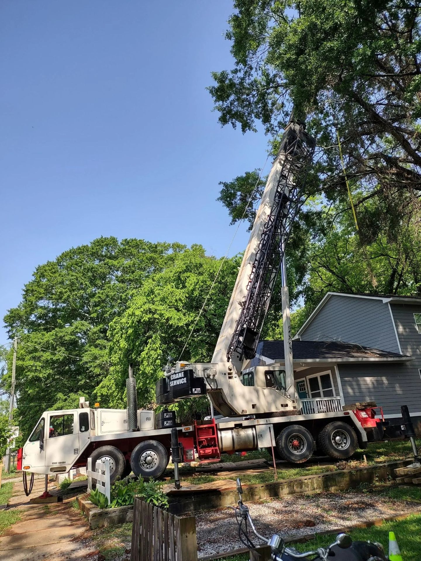 Large crane trimming tree next to a house under a clear, blue sky.