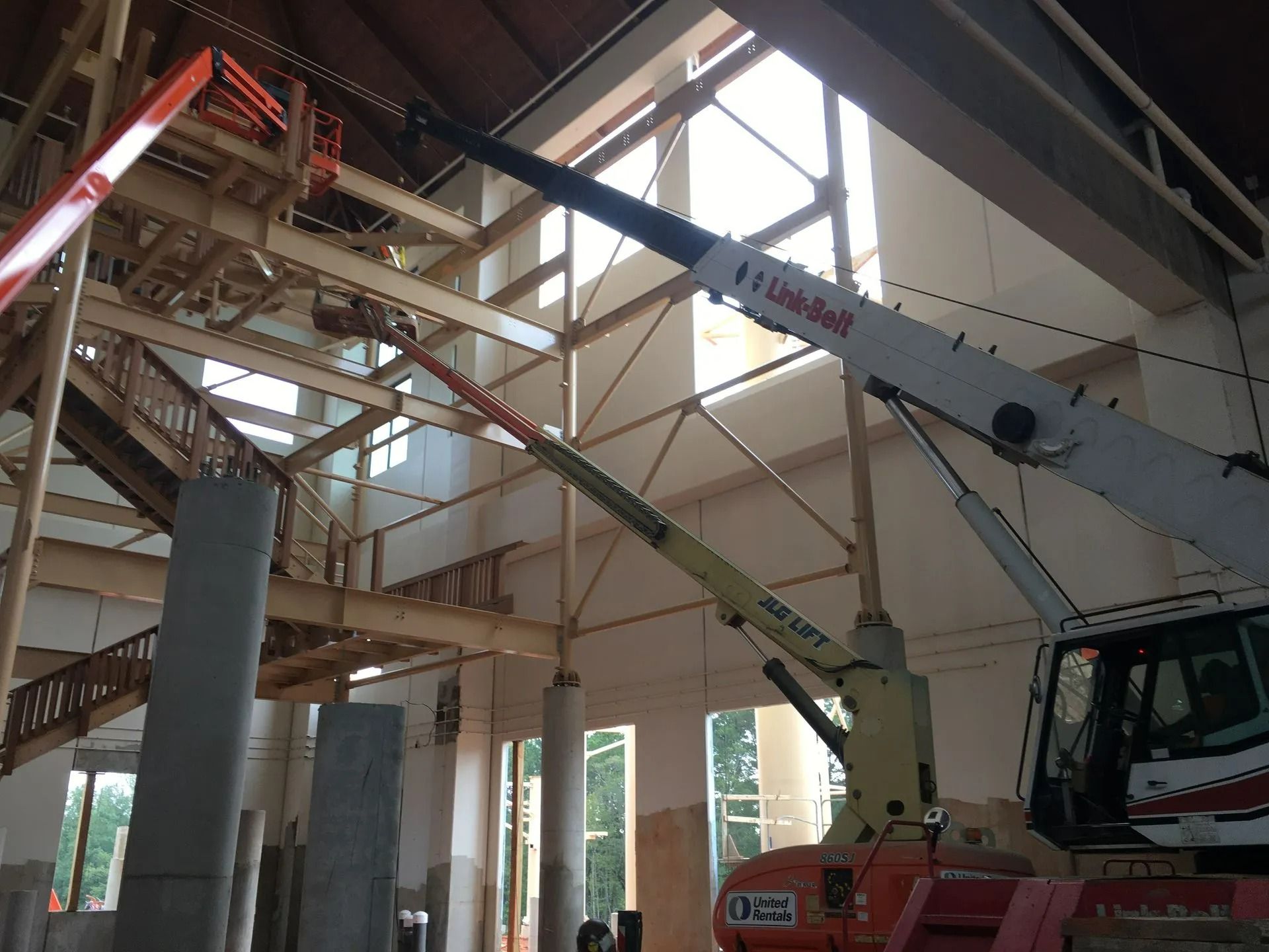 Construction site interior, boom lifts reaching up to wooden rafters. Sunlight streams through large windows.