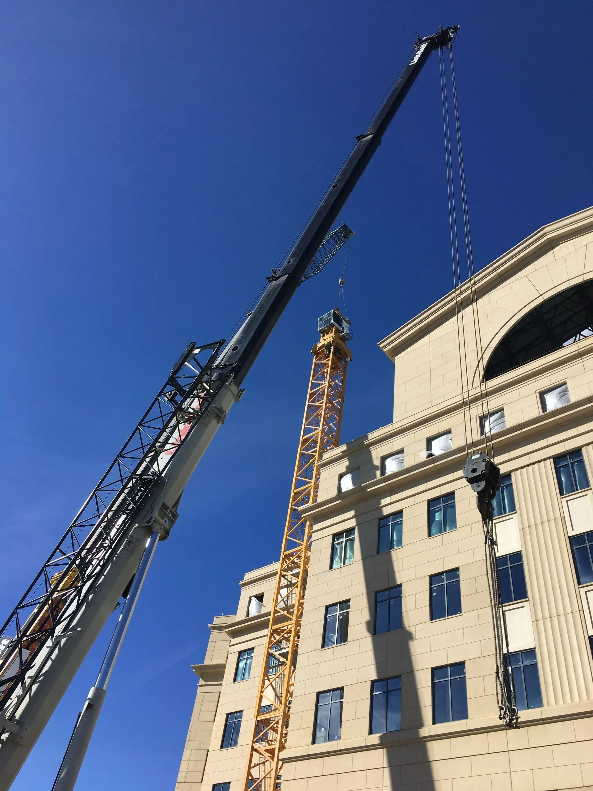 A tall crane is raising a yellow tower crane next to a multi-story building against a clear blue sky.