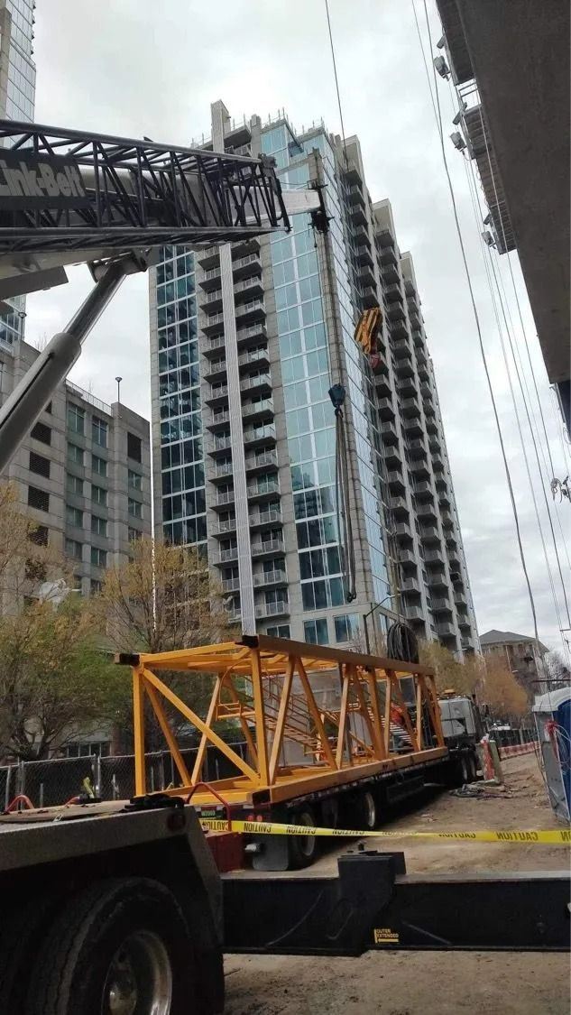 Construction site with a tall building and crane arm being delivered on a flatbed truck.