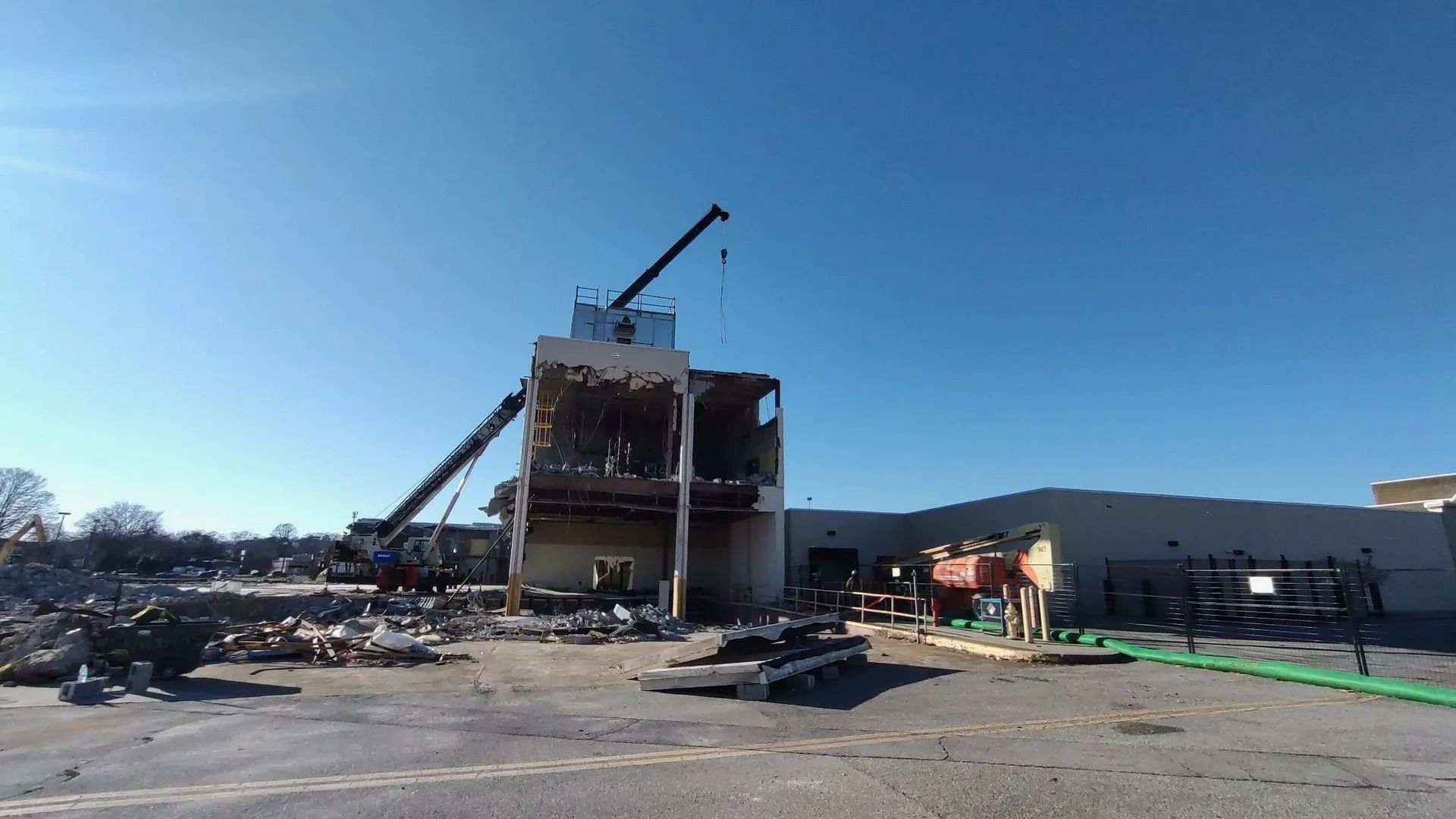 Demolition site with crane removing a building's upper structure under a blue sky. Debris and equipment surround.