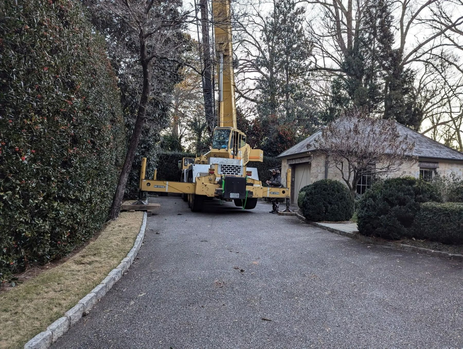 Yellow crane on driveway, trees being trimmed. Small stone building in background.