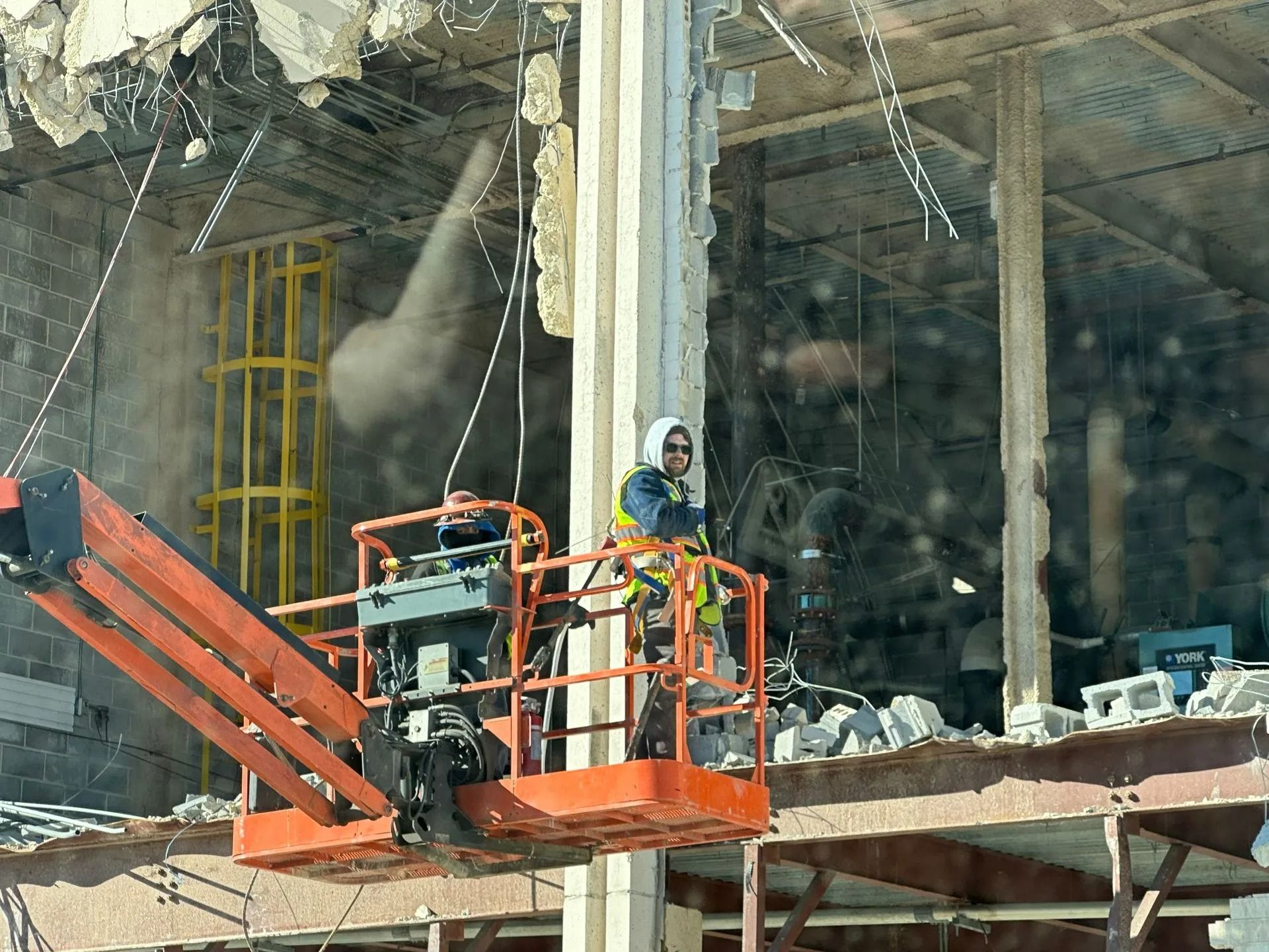 Workers in an aerial lift demolishing a building, with exposed concrete and a yellow support structure visible.