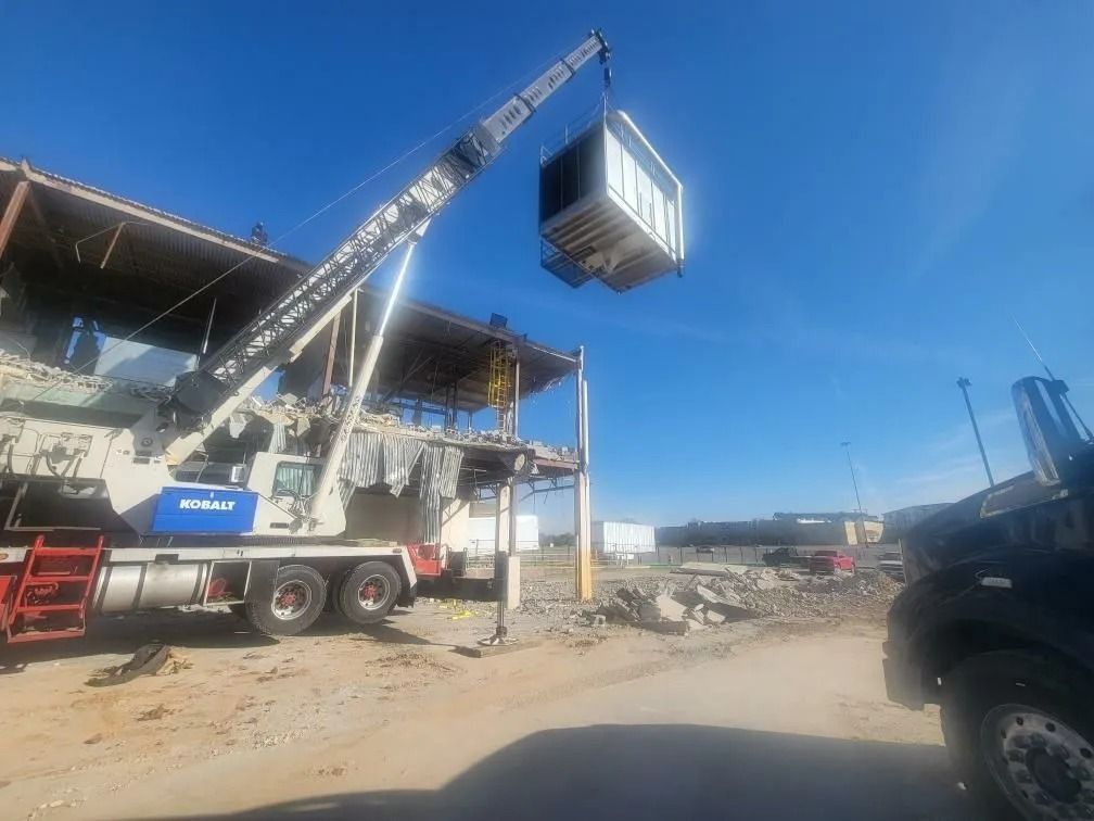 A crane lifts a large white object from a partially demolished building on a sunny day.