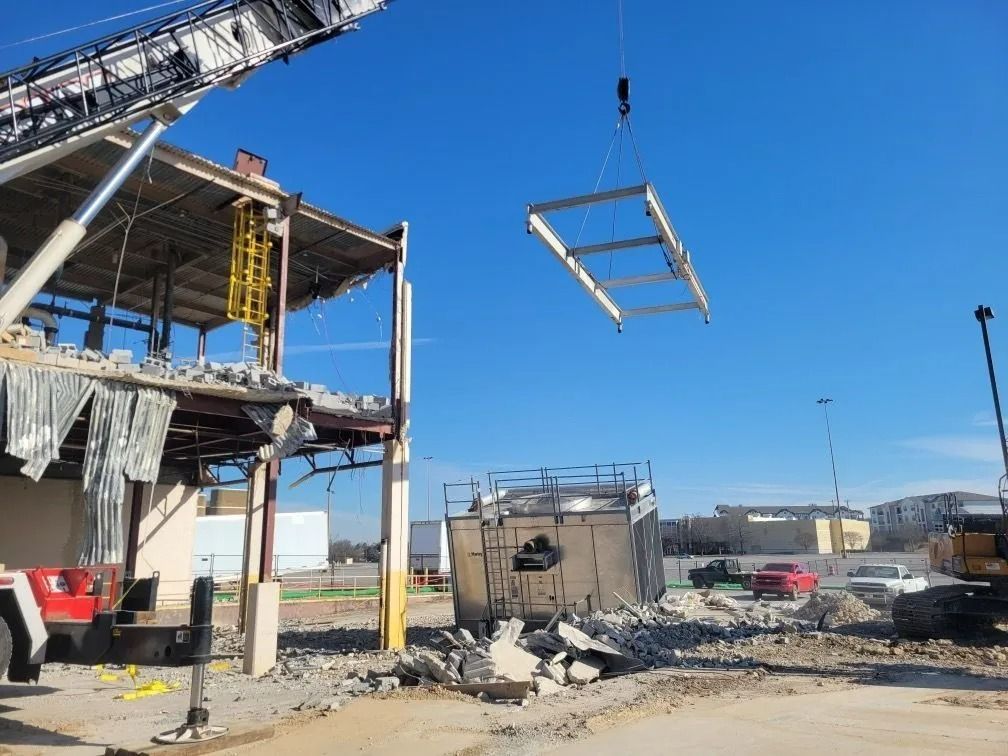A crane lifts a metal frame from a building being demolished; debris and equipment are on-site.